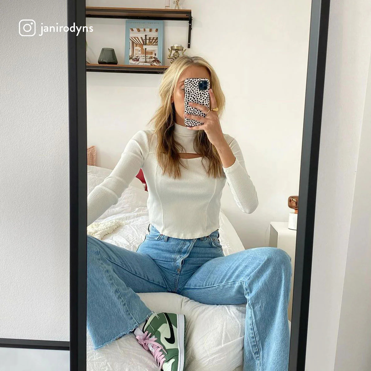 Donna allo specchio, indossa top bianco e jeans blu, sneakers verdi e nere, e telefono in mano.