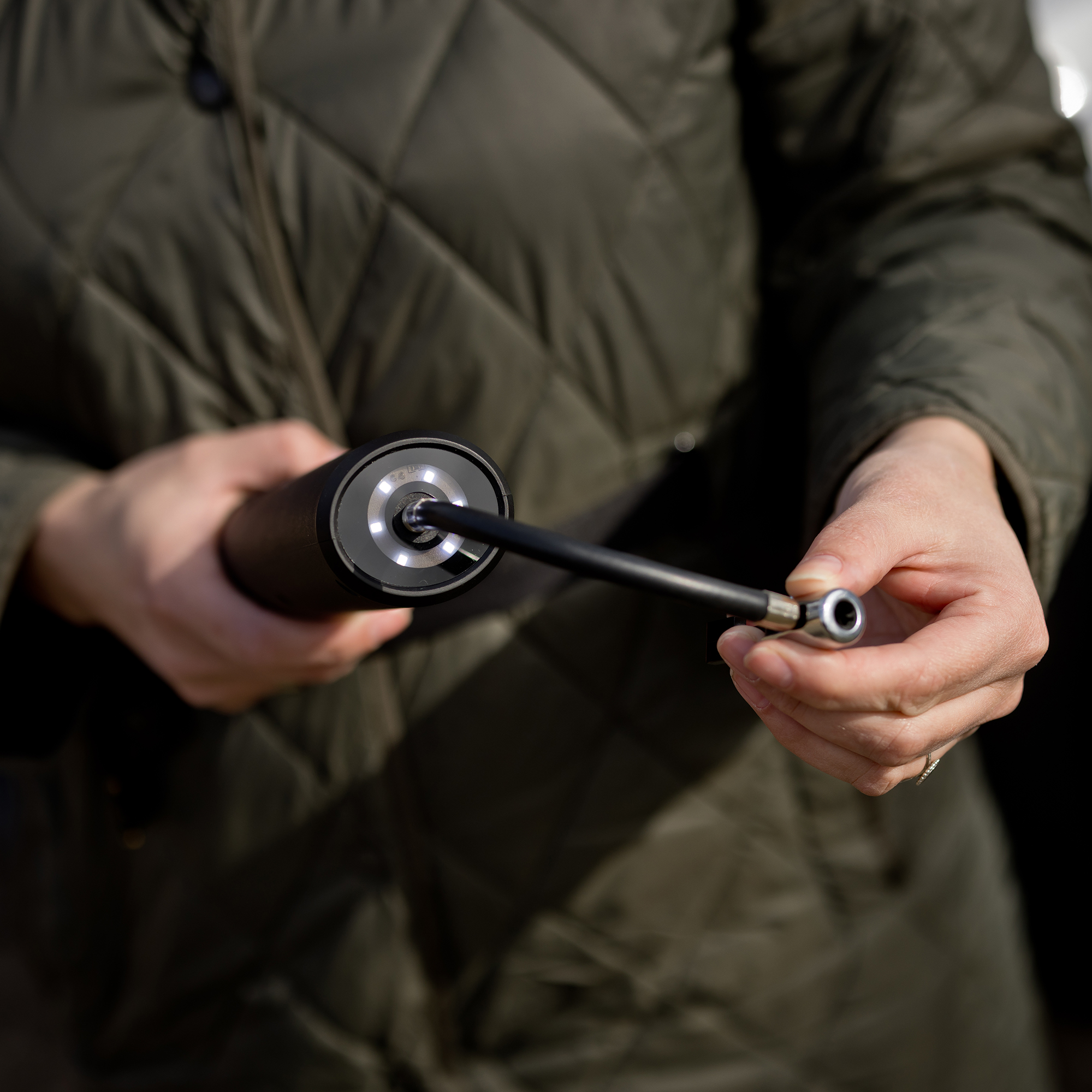 A person holding a black device with a silver nozzle. They are wearing a green jacket.
