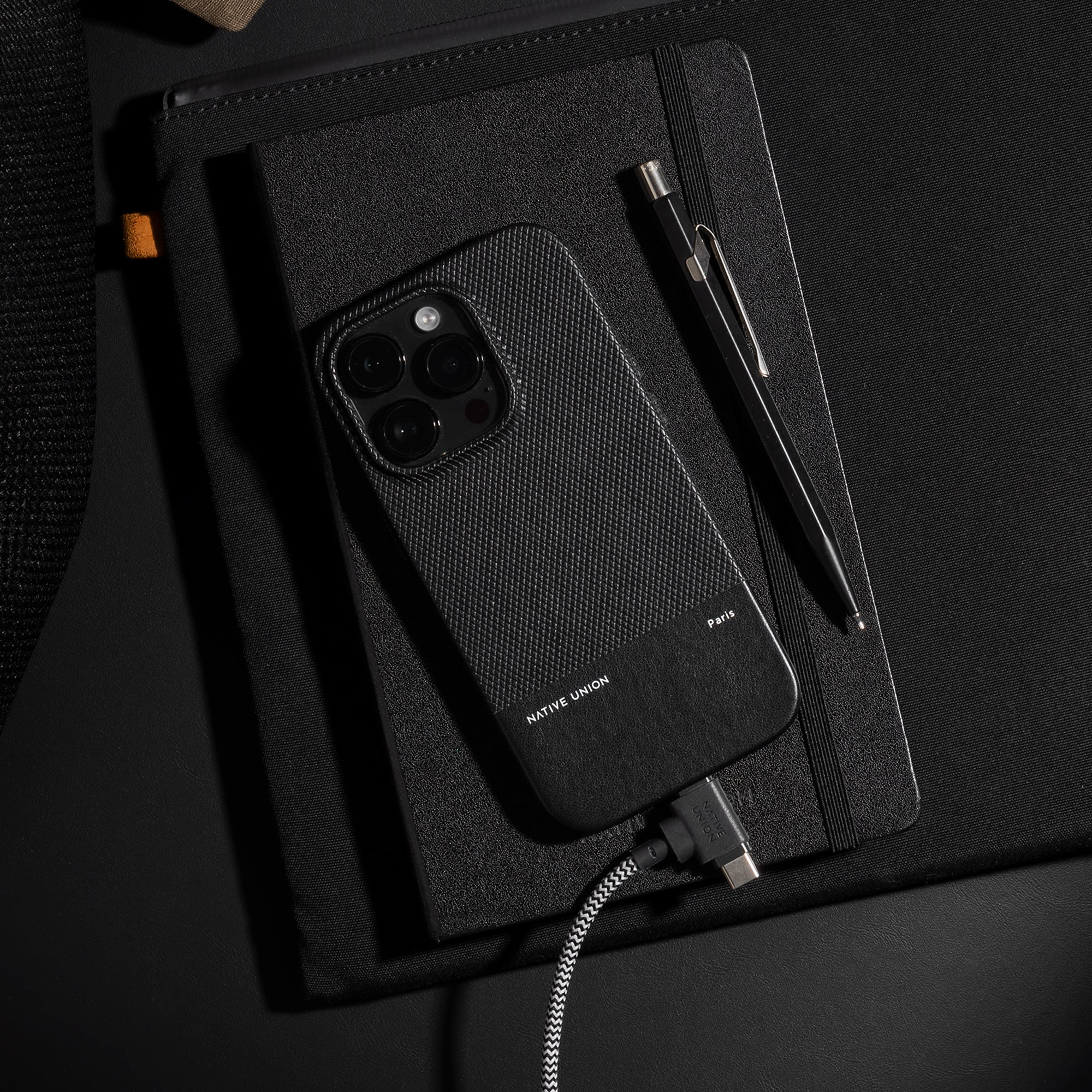 Telefono nero in custodia Native Union, in carica con cavo USB-C su un taccuino.