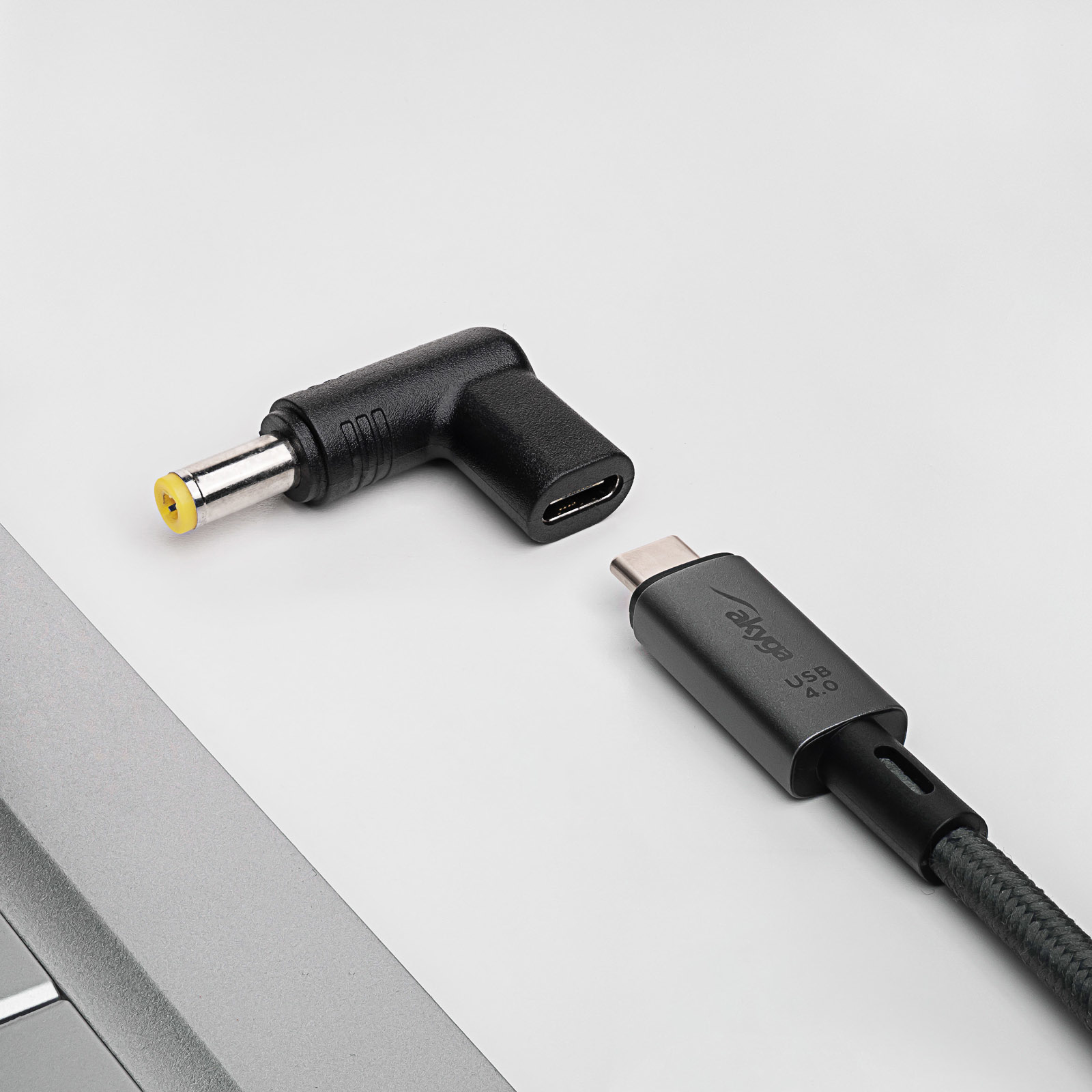 Un adattatore nero a L e un cavo USB-C nero, vicino a un laptop, sfondo bianco.