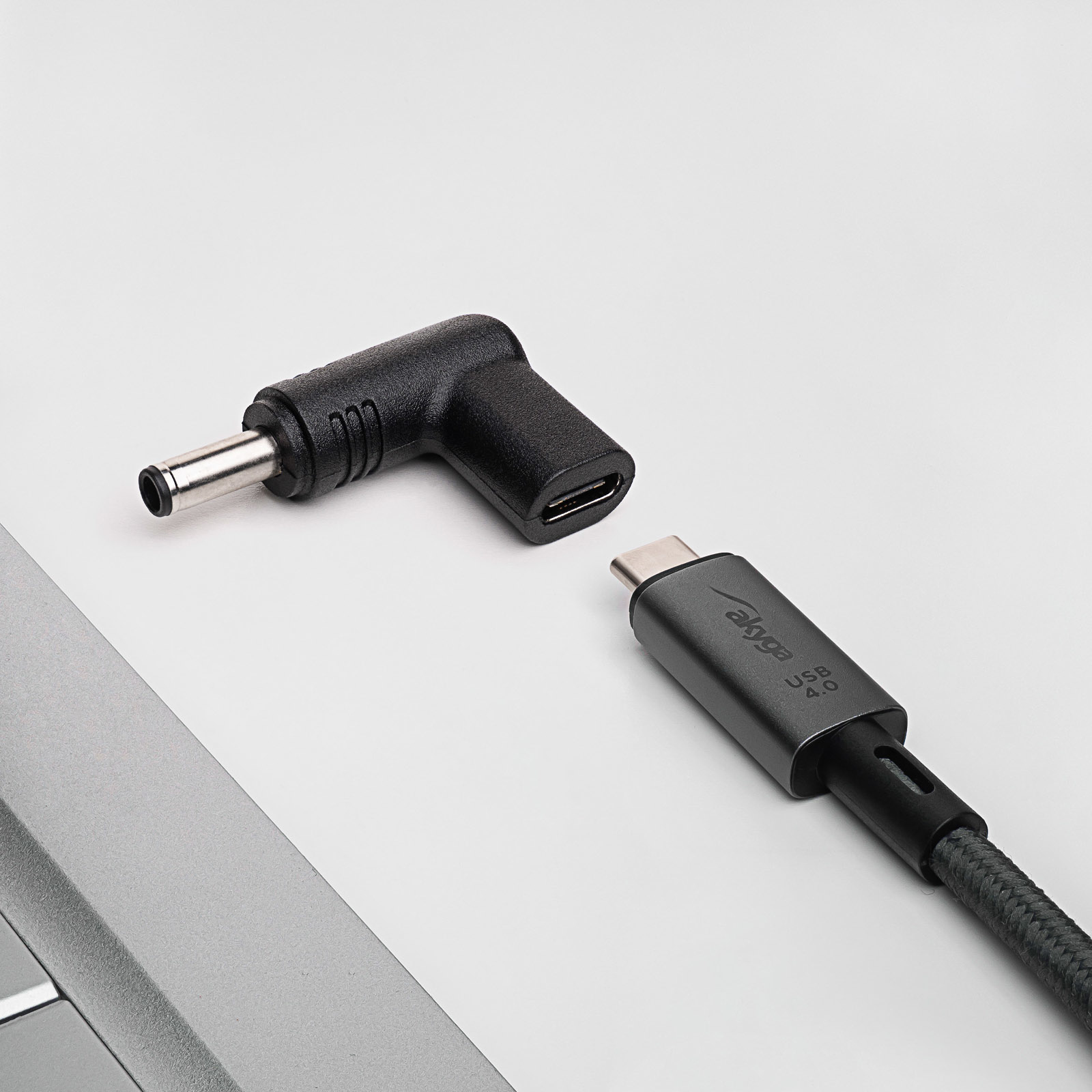 Un adattatore nero a L e un cavo USB-C nero, vicino a un laptop, sfondo bianco.