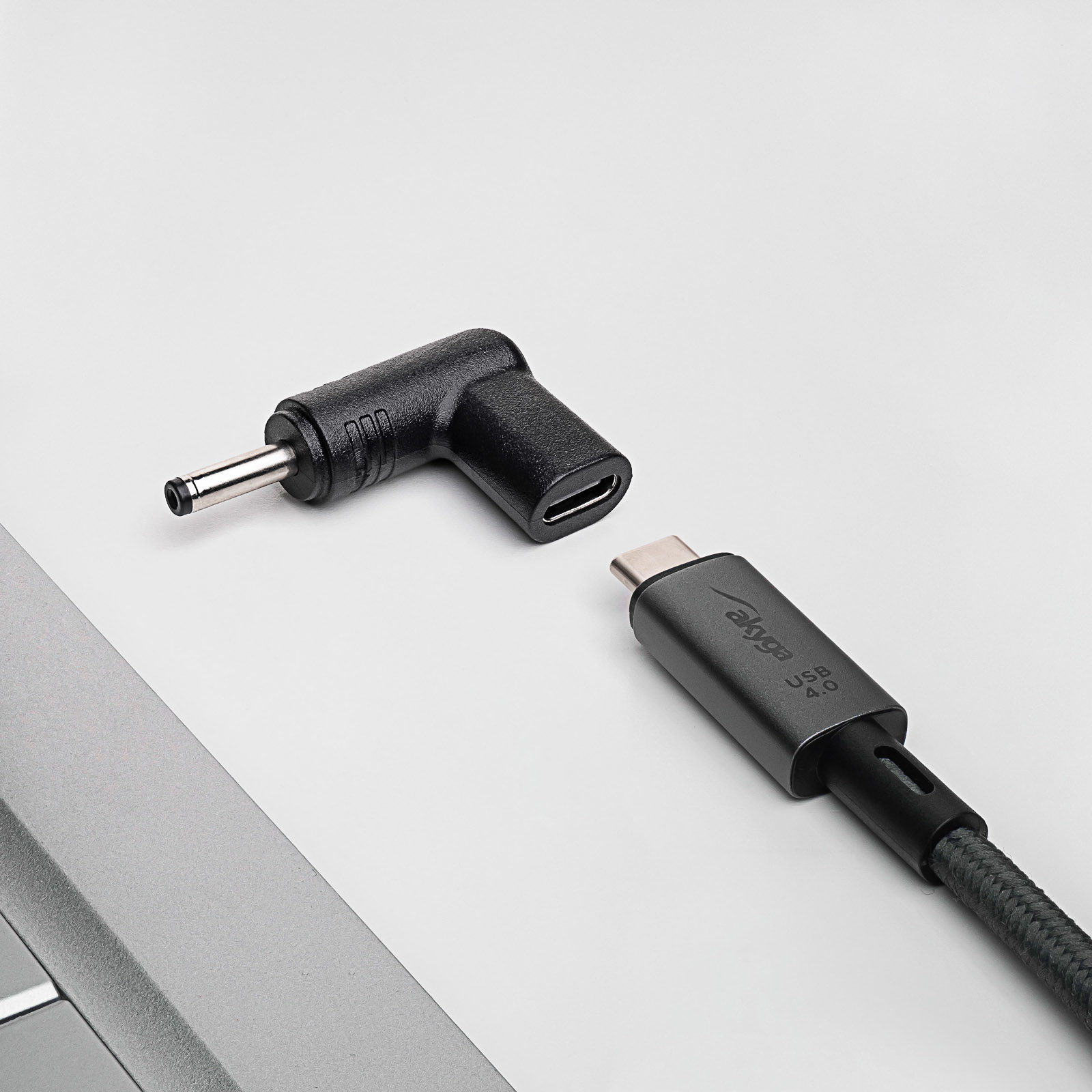 Adattatore nero e cavo USB-C su superficie bianca vicino a un laptop.