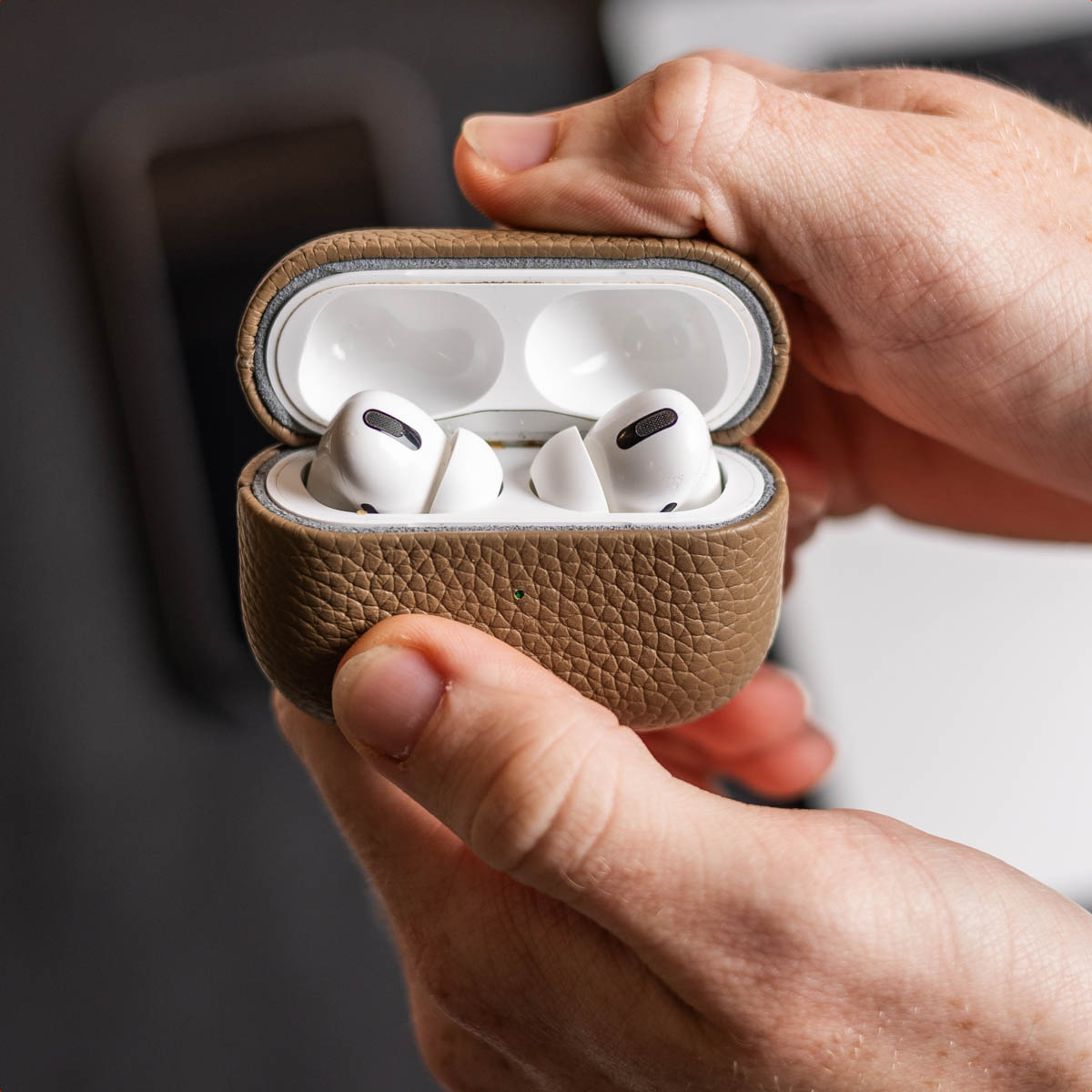 Hands holding an open, brown leather case with white earbuds. The background is blurred.