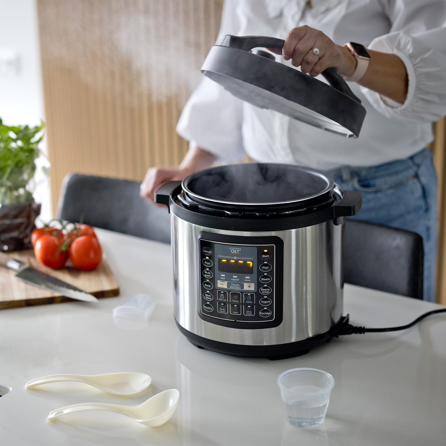 A woman opening a stainless steel pressure cooker. Steam rises. Tomatoes and utensils on counter.