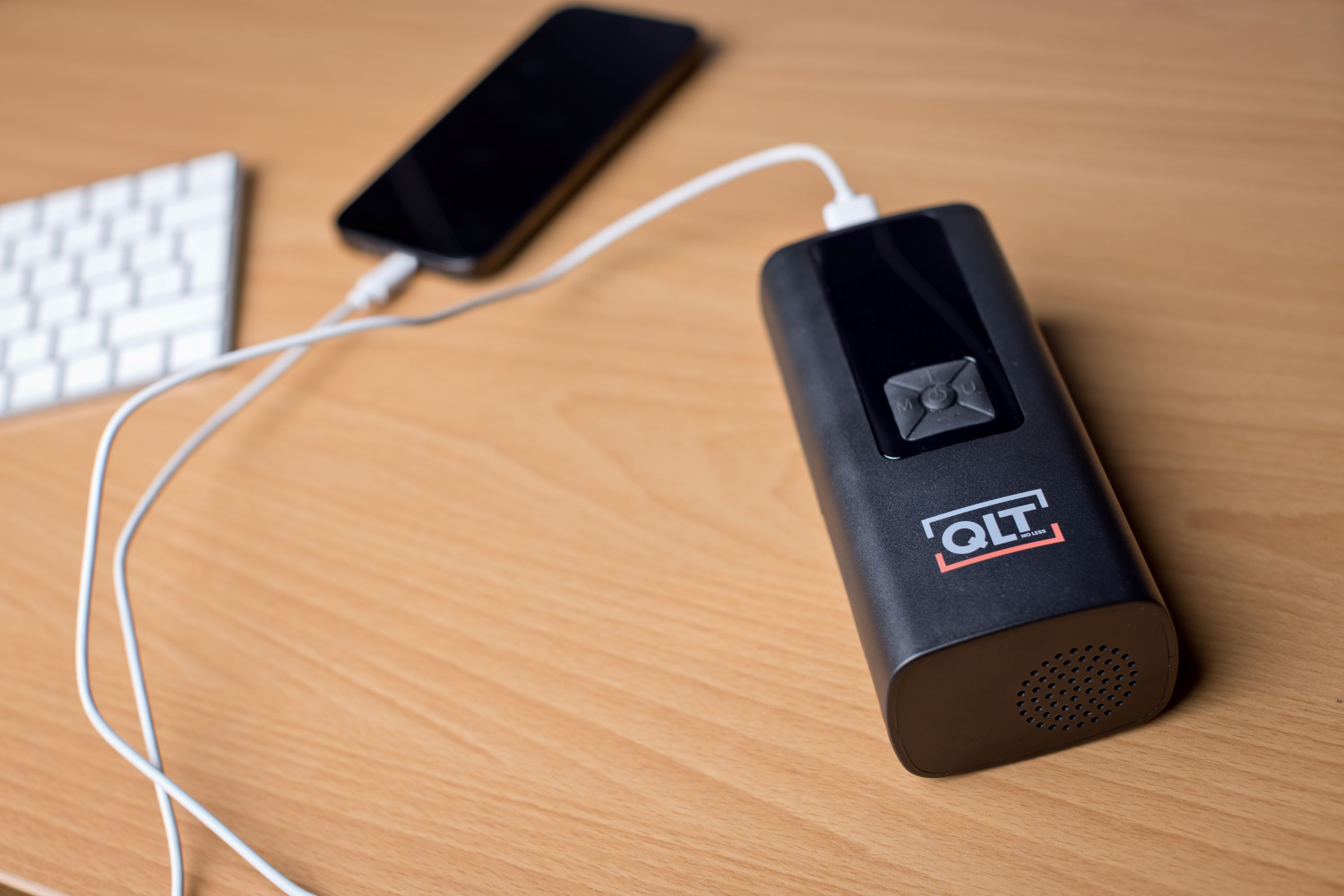 A black power bank is charging a phone. A white keyboard is in the background on a wooden surface.