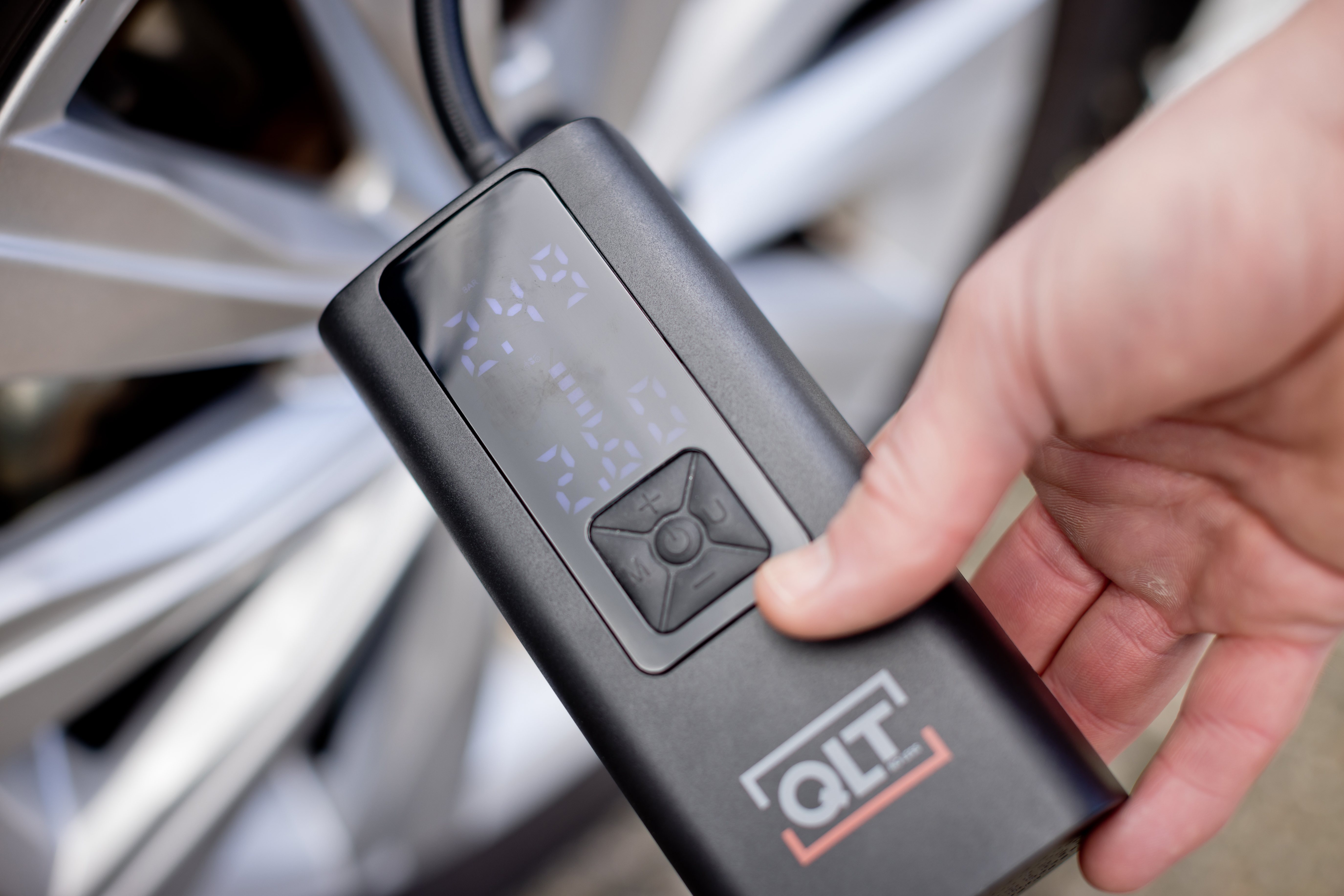 Hand holding a black QLT tire inflator, showing pressure reading, with a car wheel in the background.