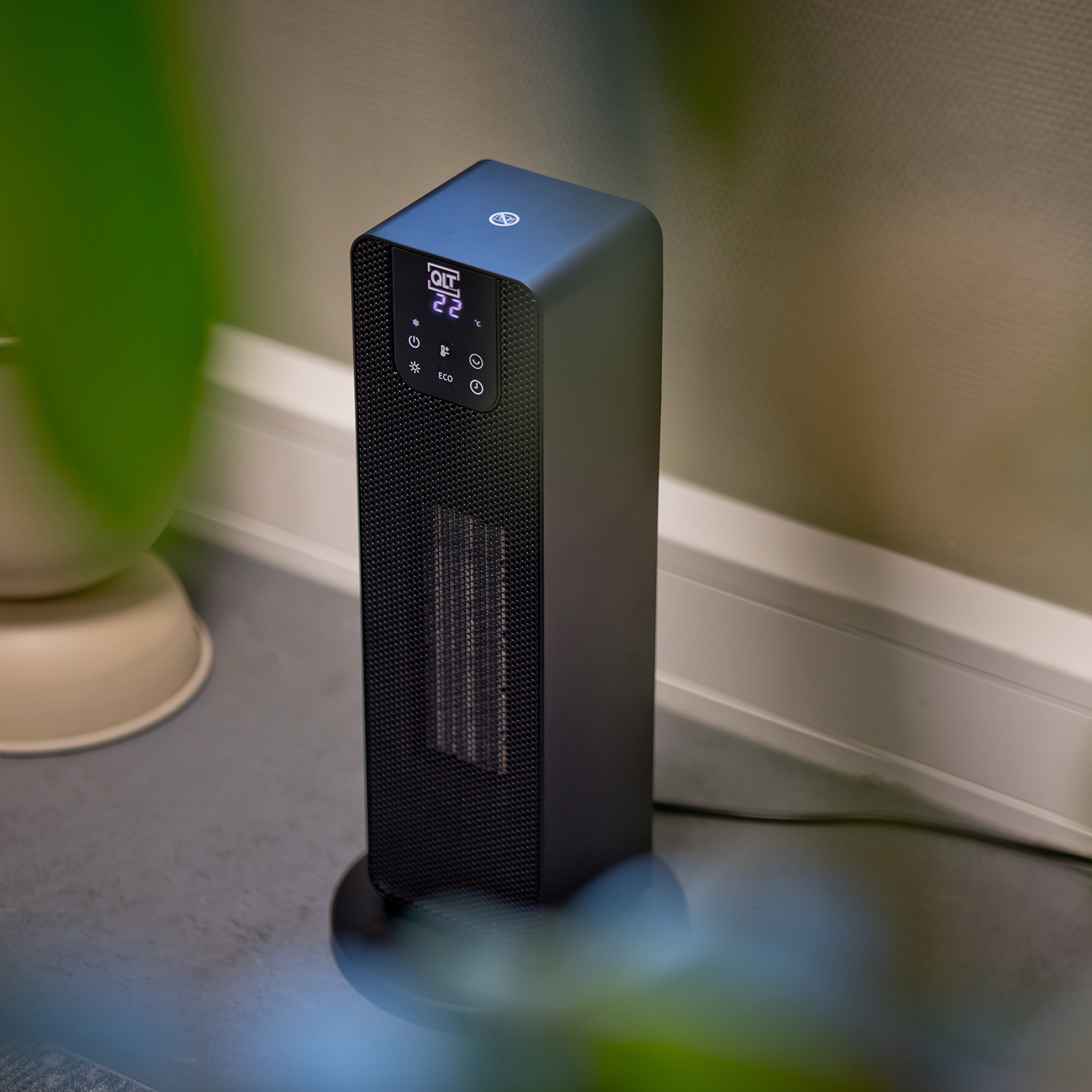 A black, vertical electric heater on a gray floor, with a green plant in the foreground.