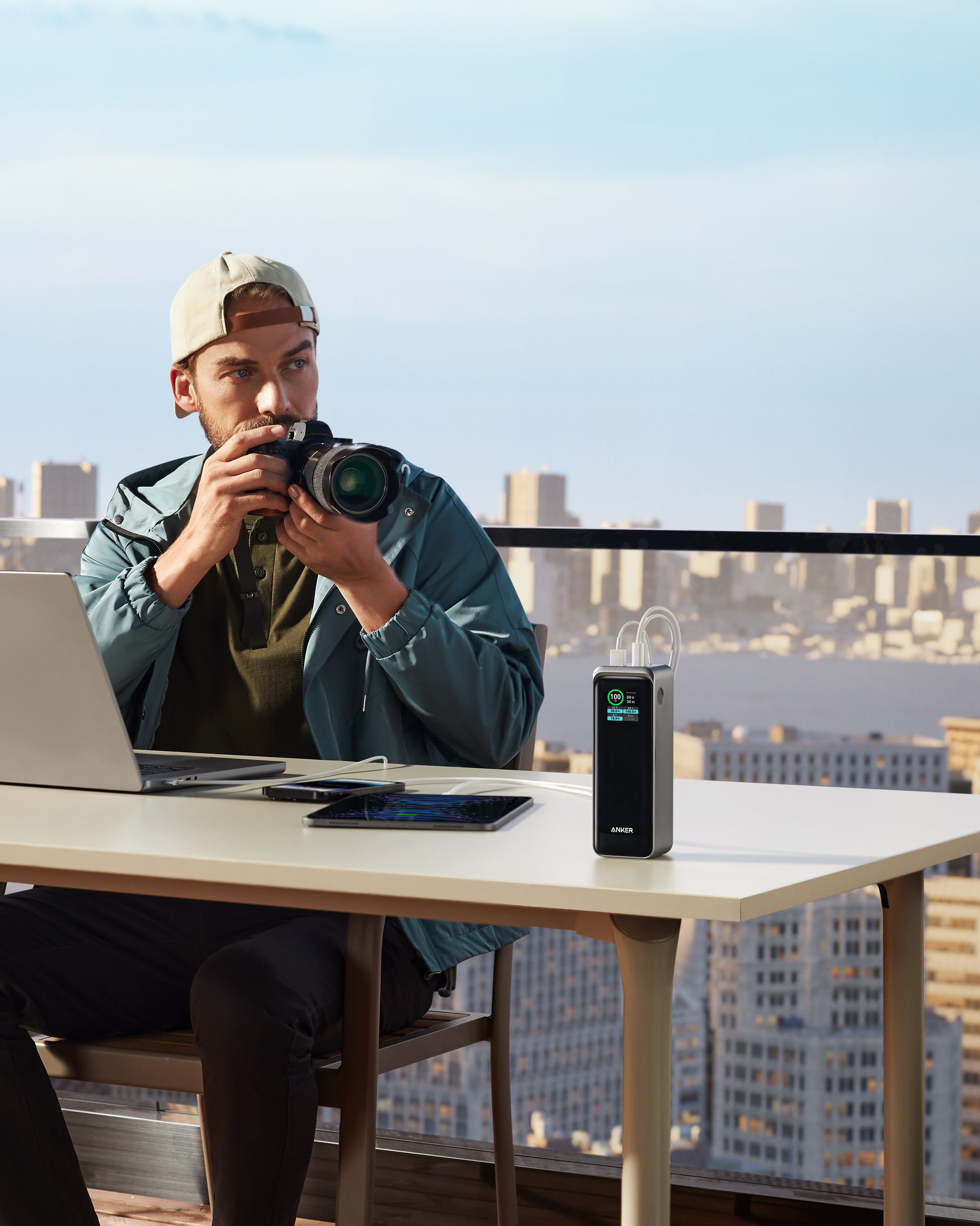 Un uomo scatta una foto con una fotocamera, accanto a un laptop e a un power bank. Città sullo sfondo.