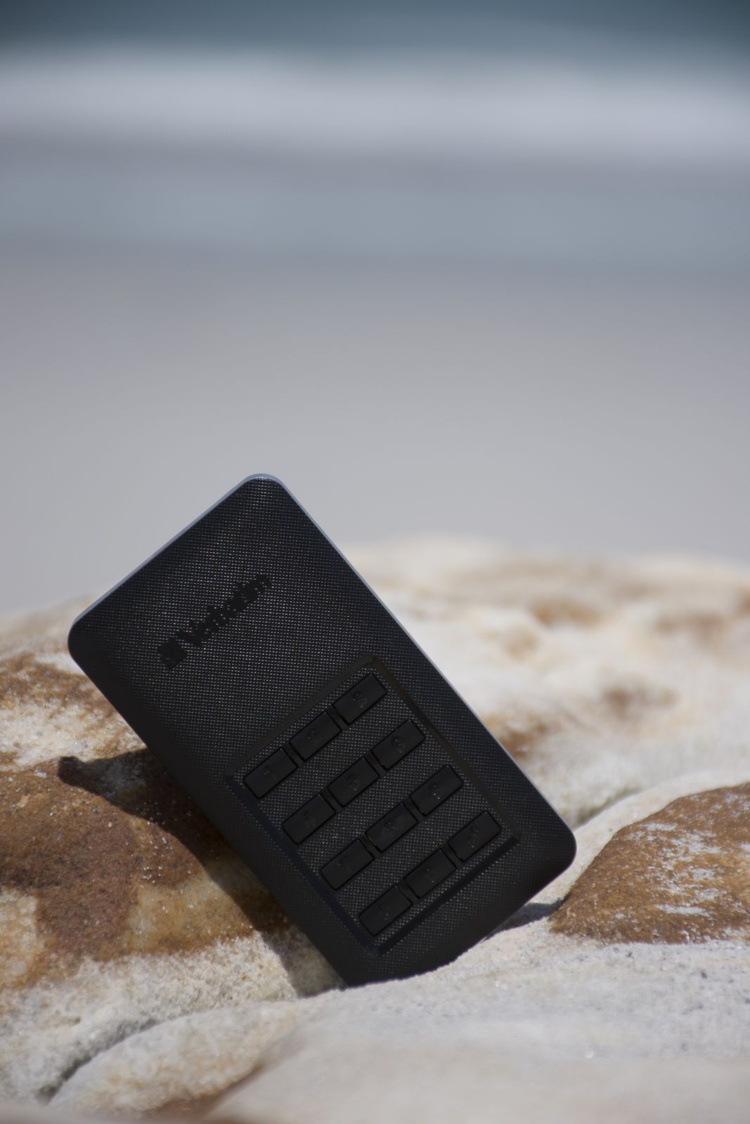 Czarne urządzenie z przyciskami na skale, tło plaży. Urządzenie leży po przekątnej.