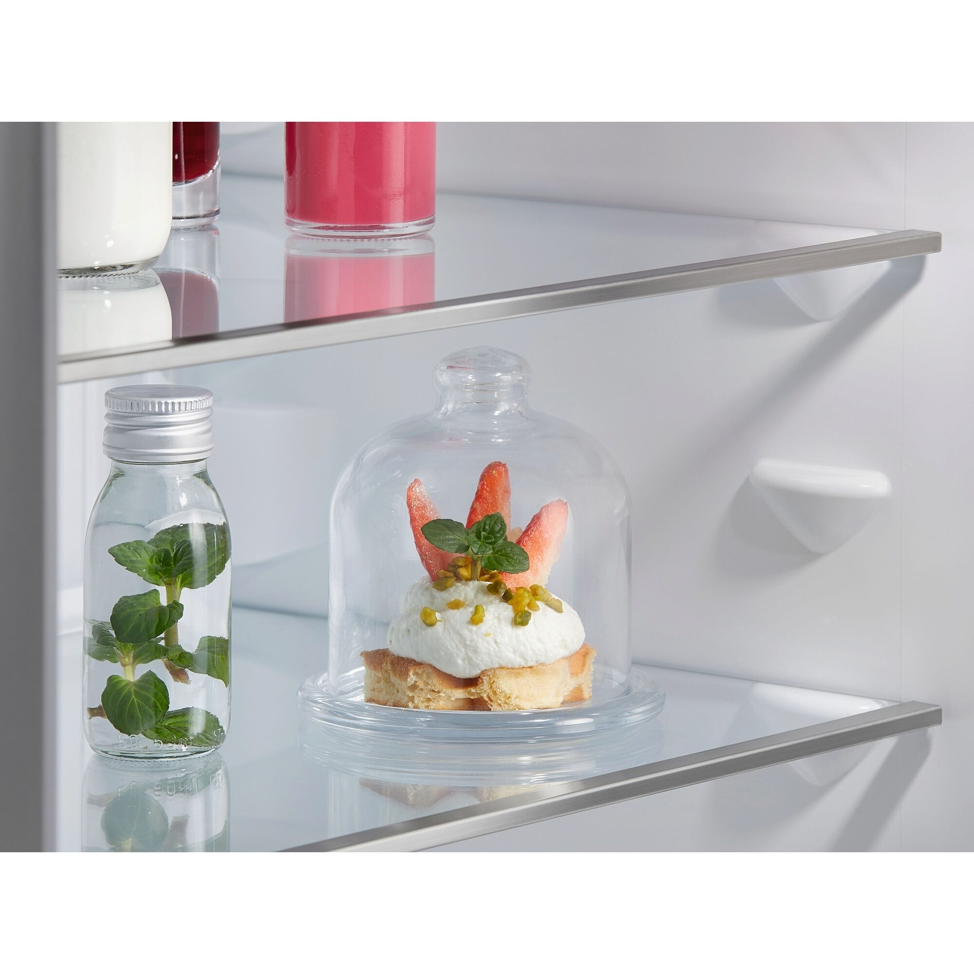 Una campana de cristal sobre un postre en una estantería de cristal. Otros artículos como una botella también están en la estantería.