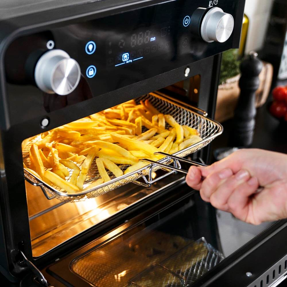 Frytownica powietrzna z frytkami w środku. Ręka sięga do frytownicy powietrznej z obcęgami.