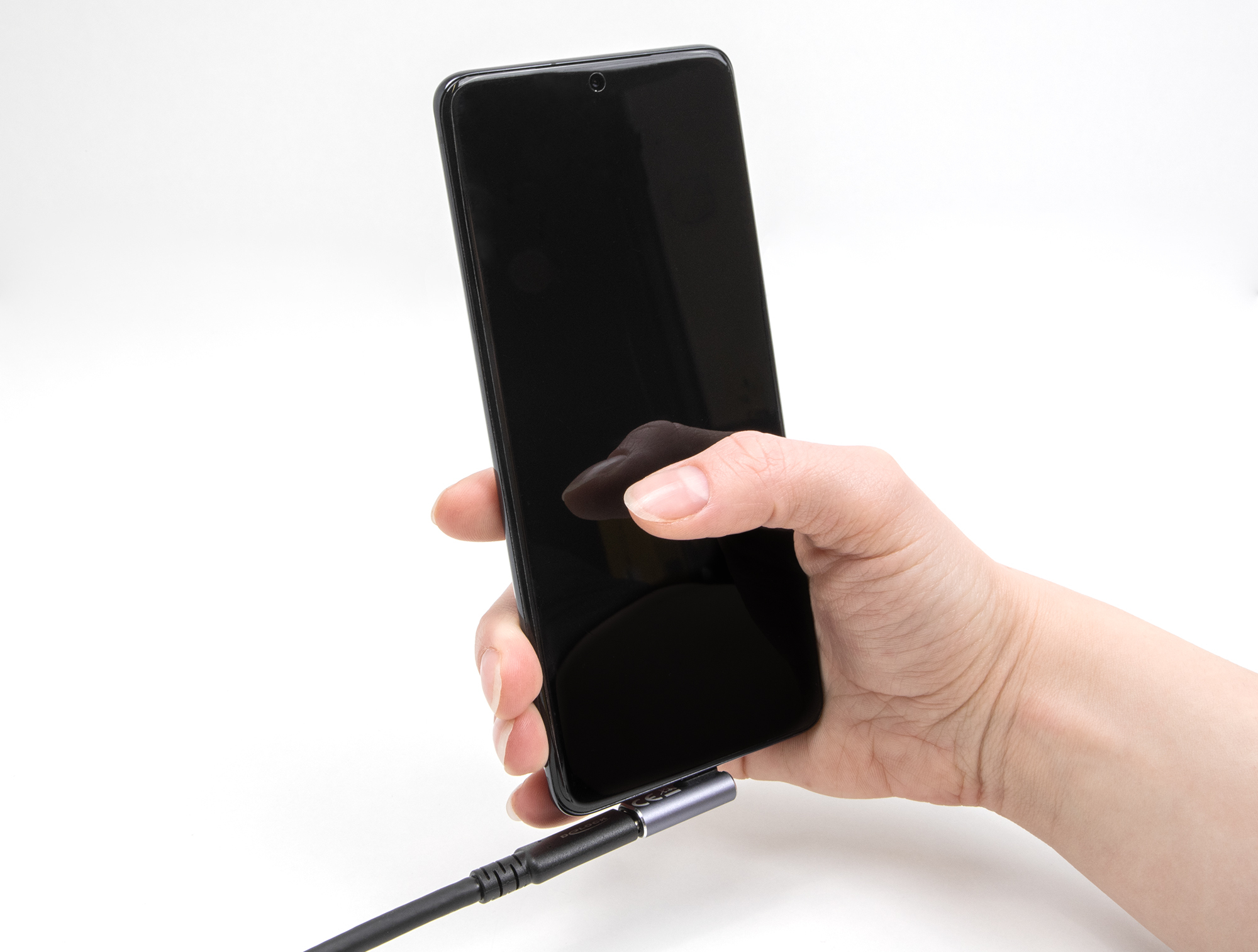 Una mano che tiene uno smartphone nero, collegato a un cavo USB-C nero. Sfondo bianco.