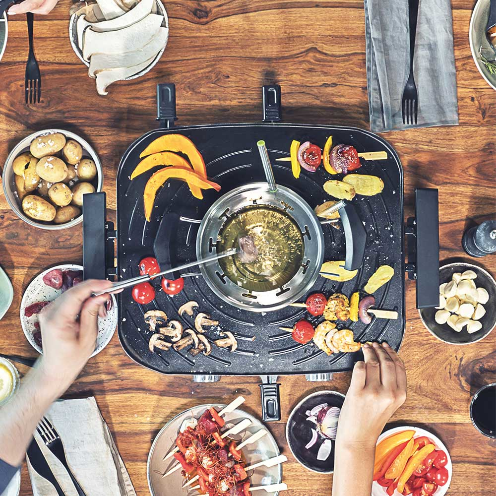 Tisch mit Essen und Händen: Fonduetopf, Gemüsespieße, Kartoffeln und Teller mit Essen.