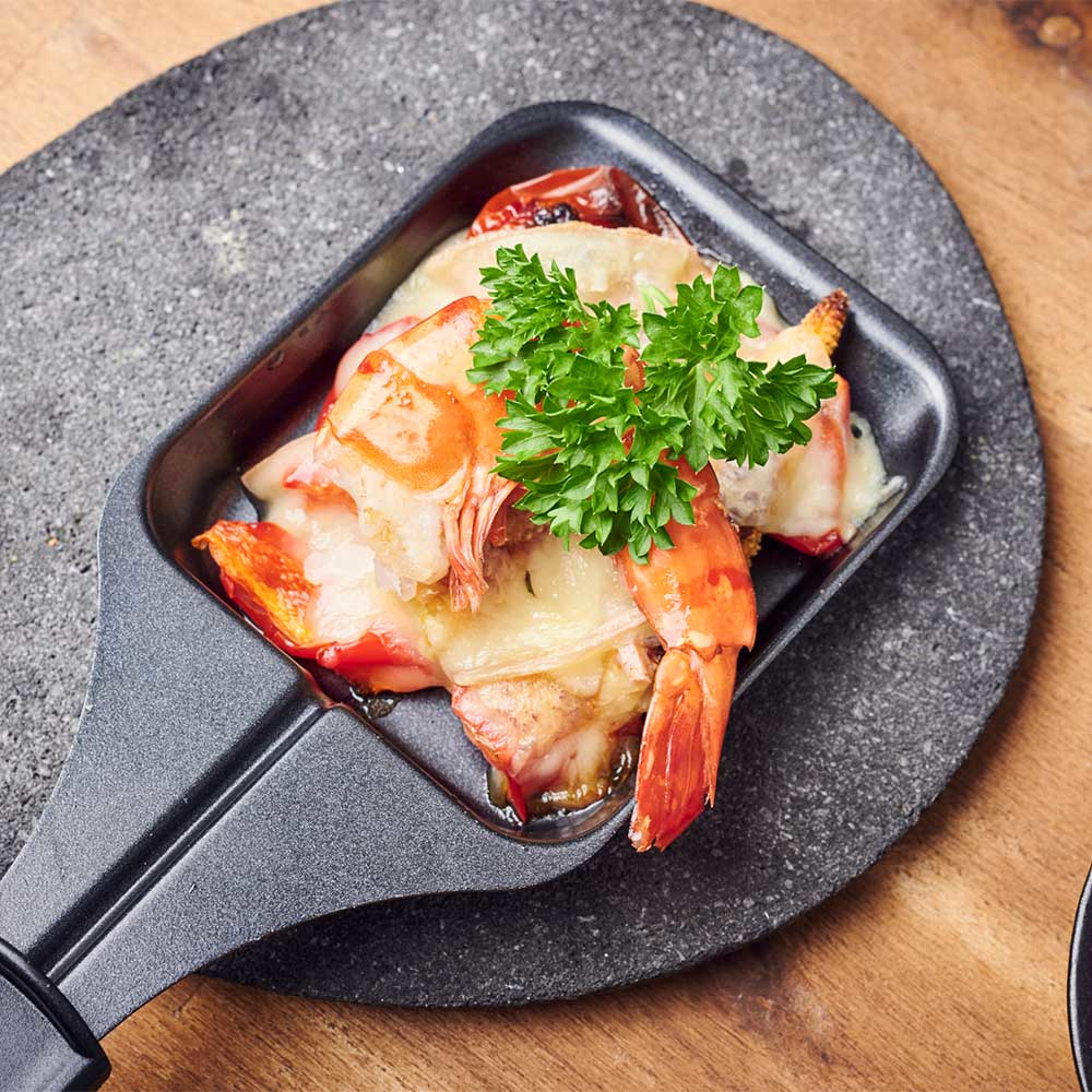 Eine kleine, schwarze Raclette-Pfanne mit Garnelen und Käse, garniert mit Petersilie.