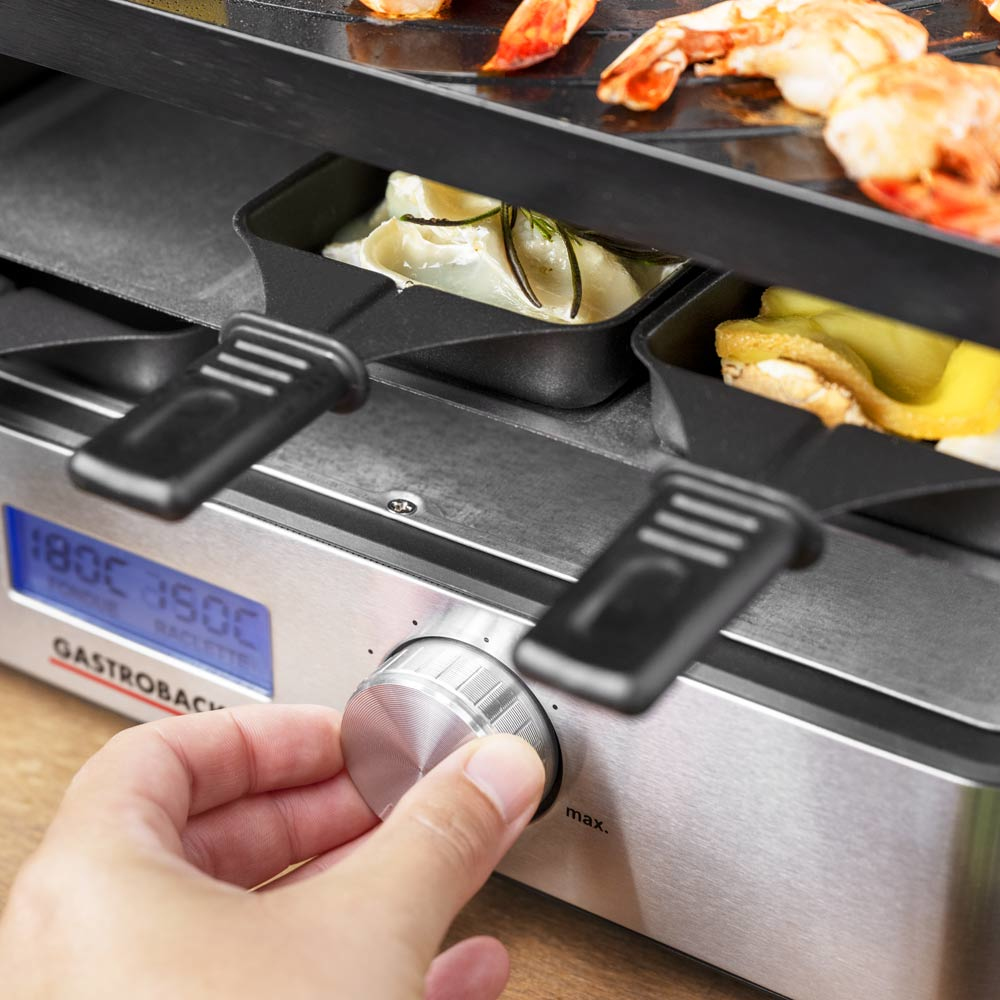 Una mano che gira una manopola su una macchina per raclette Gastrobac con cibo nei vassoi.