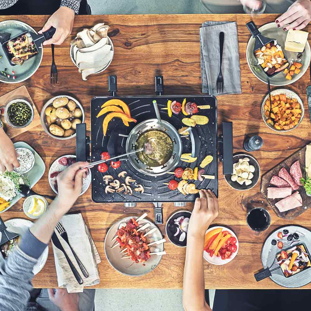 Persone attorno a un tavolo in legno con una griglia per raclette. Cibo sui piatti. Mani che raggiungono il cibo.