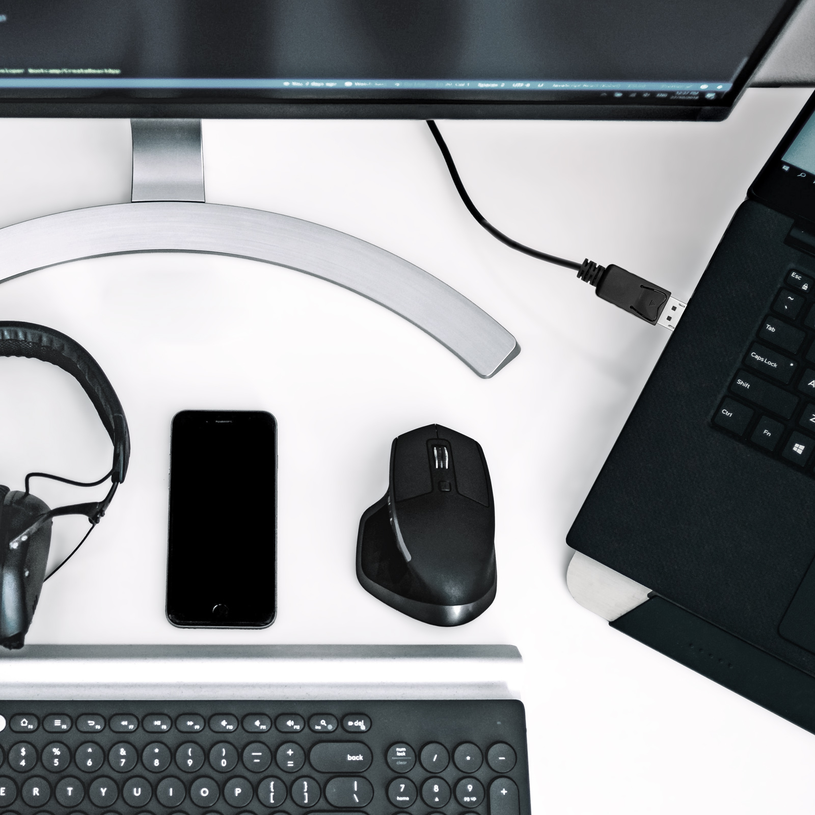 Konfiguracja biurka: monitor, słuchawki, telefon, mysz, klawiatura i laptop. Wszystko na białej powierzchni.