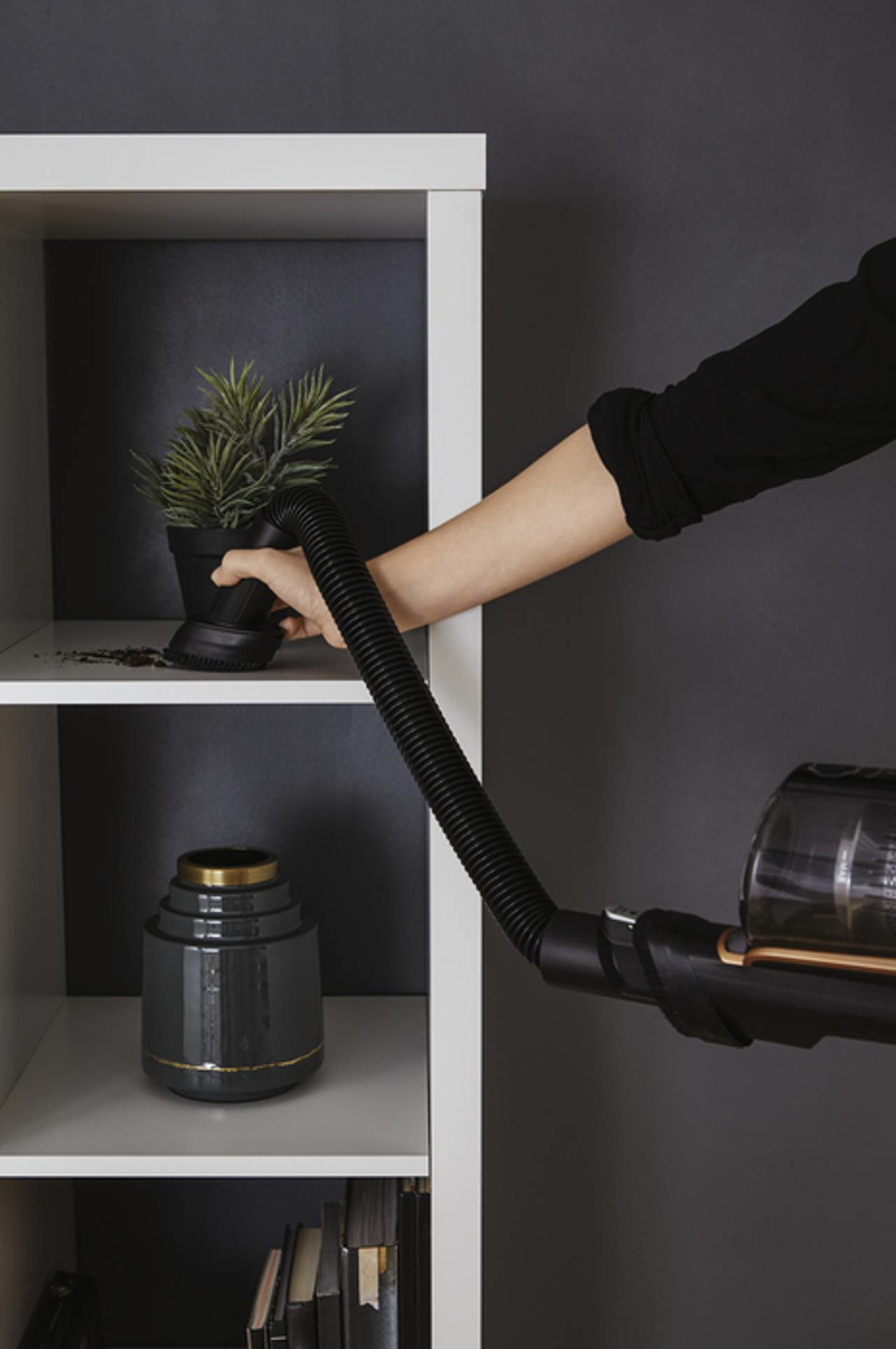 Ręka używająca odkurzacza na półce z rośliną i wazonem. Czarne i białe kolory.