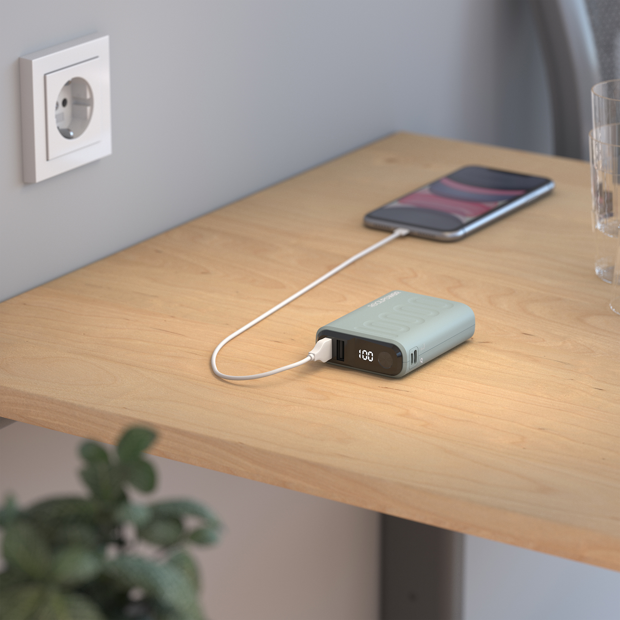 Power bank che carica un telefono su una superficie in legno, vicino a una presa a muro. Carica al 100% visualizzata.