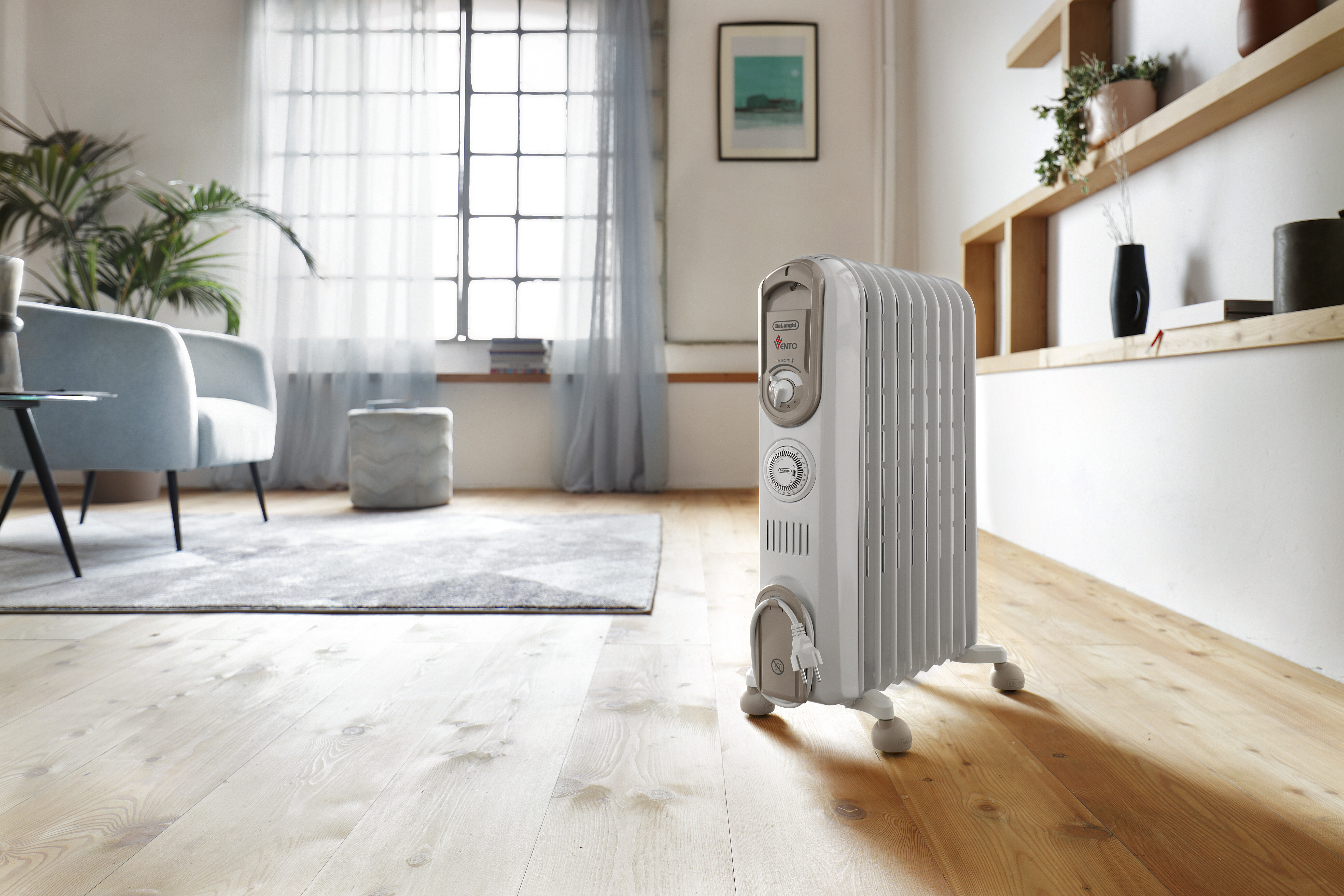 Biały grzejnik olejowy z pokrętłem i uchwytem, na drewnianej podłodze. Widoczny jasnoniebieski fotel.