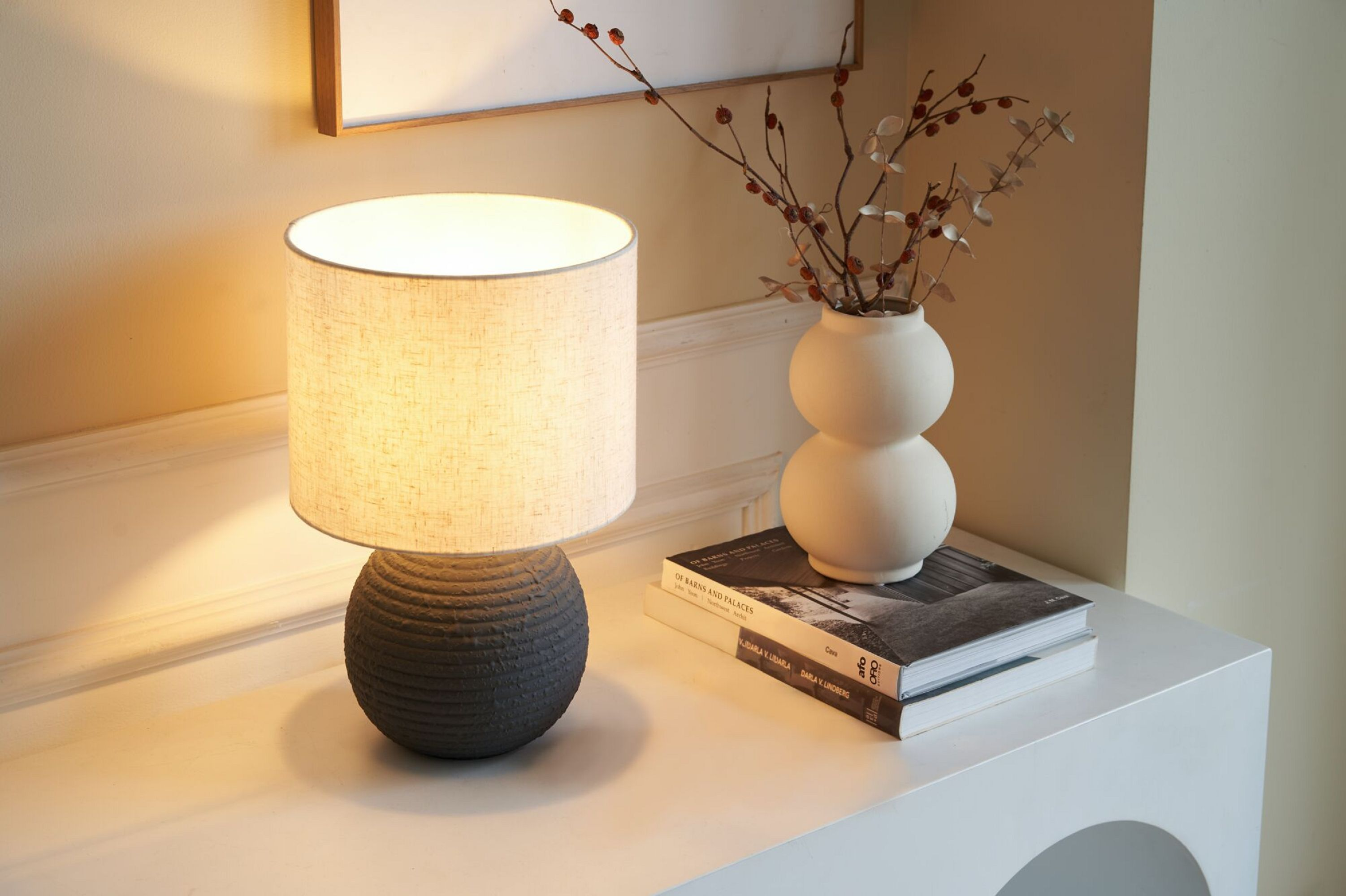 Lampa stołowa z teksturowaną podstawą, wazonem, książkami i dekoracyjnymi gałęziami.