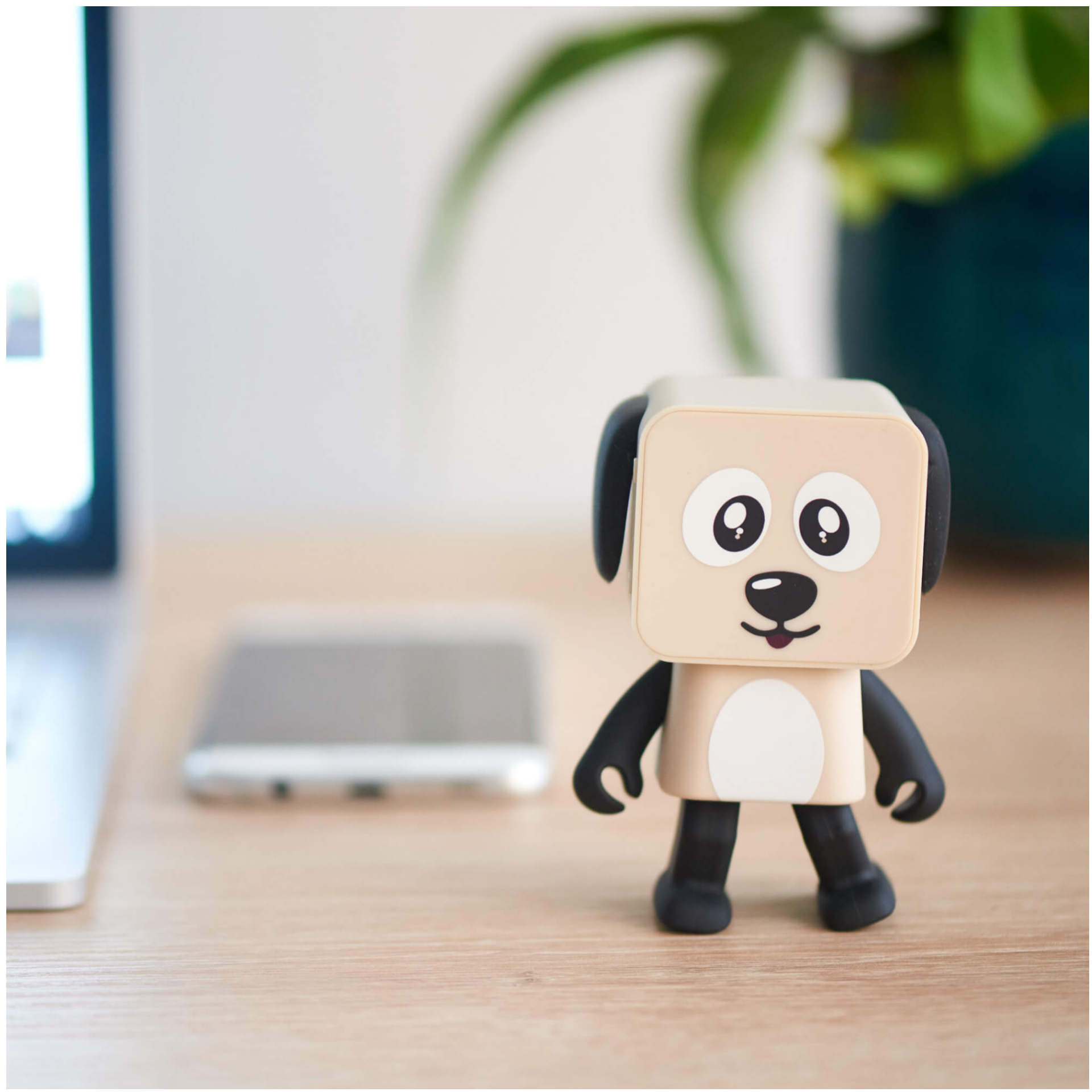 Głośnik w kształcie psa na drewnianym biurku. Telefon i laptop w tle. Biała ściana.