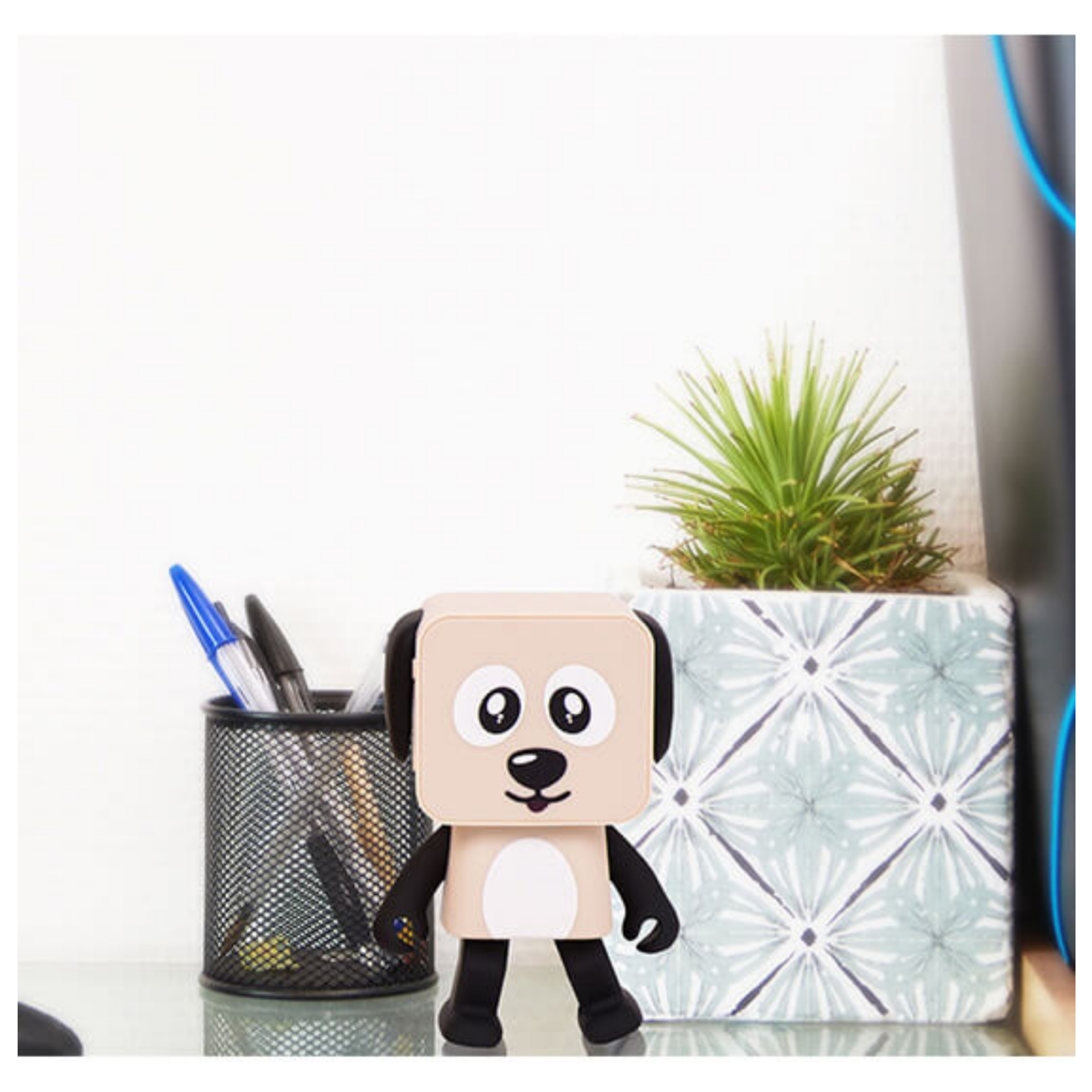 Głośnik w kształcie psa na szklanej powierzchni. Pojemnik na długopisy i roślina w tle. Biała ściana w tle.