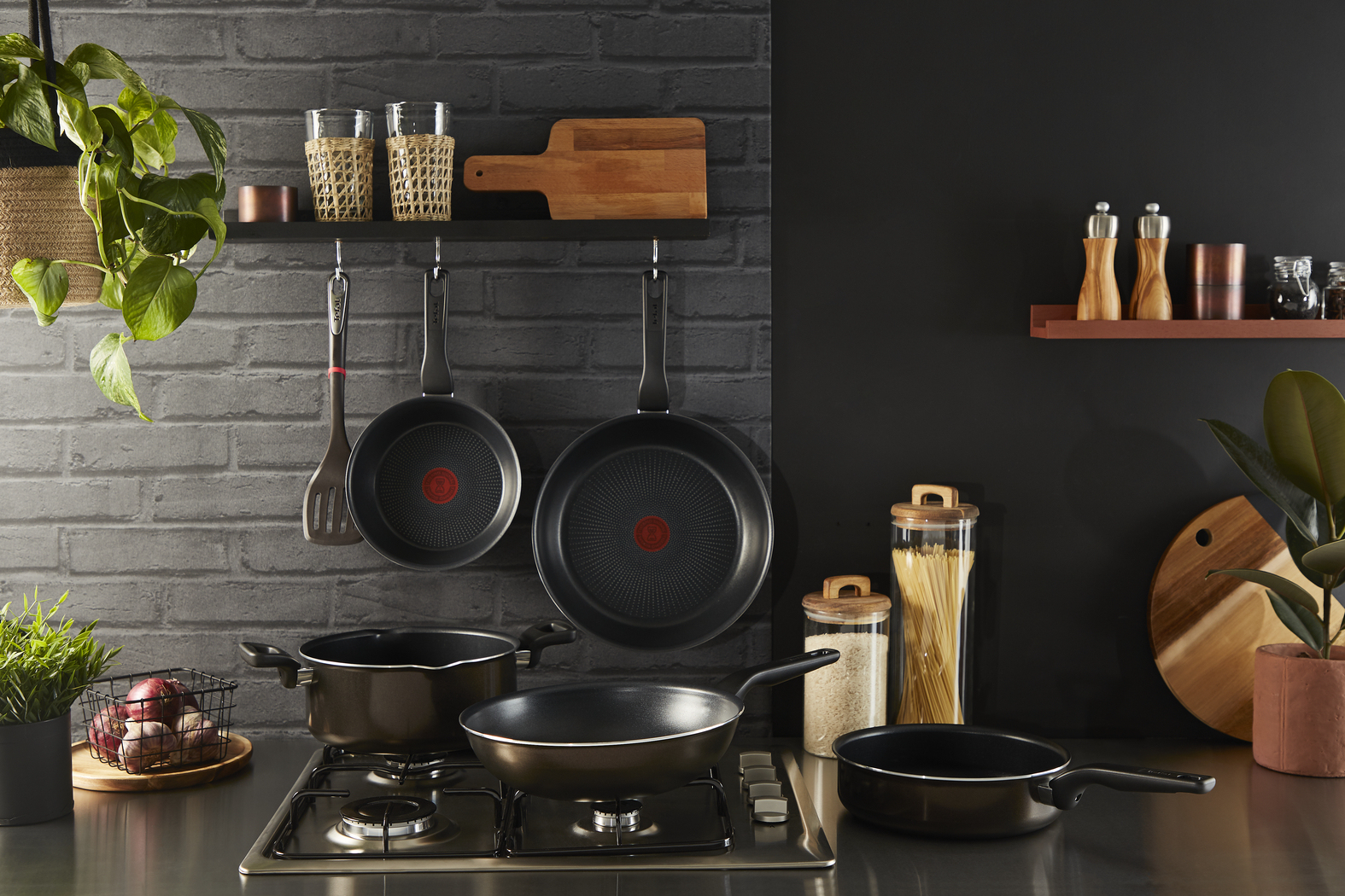Scena cucina con pentole, padelle e utensili appesi a un muro di mattoni, con fornello e piante.