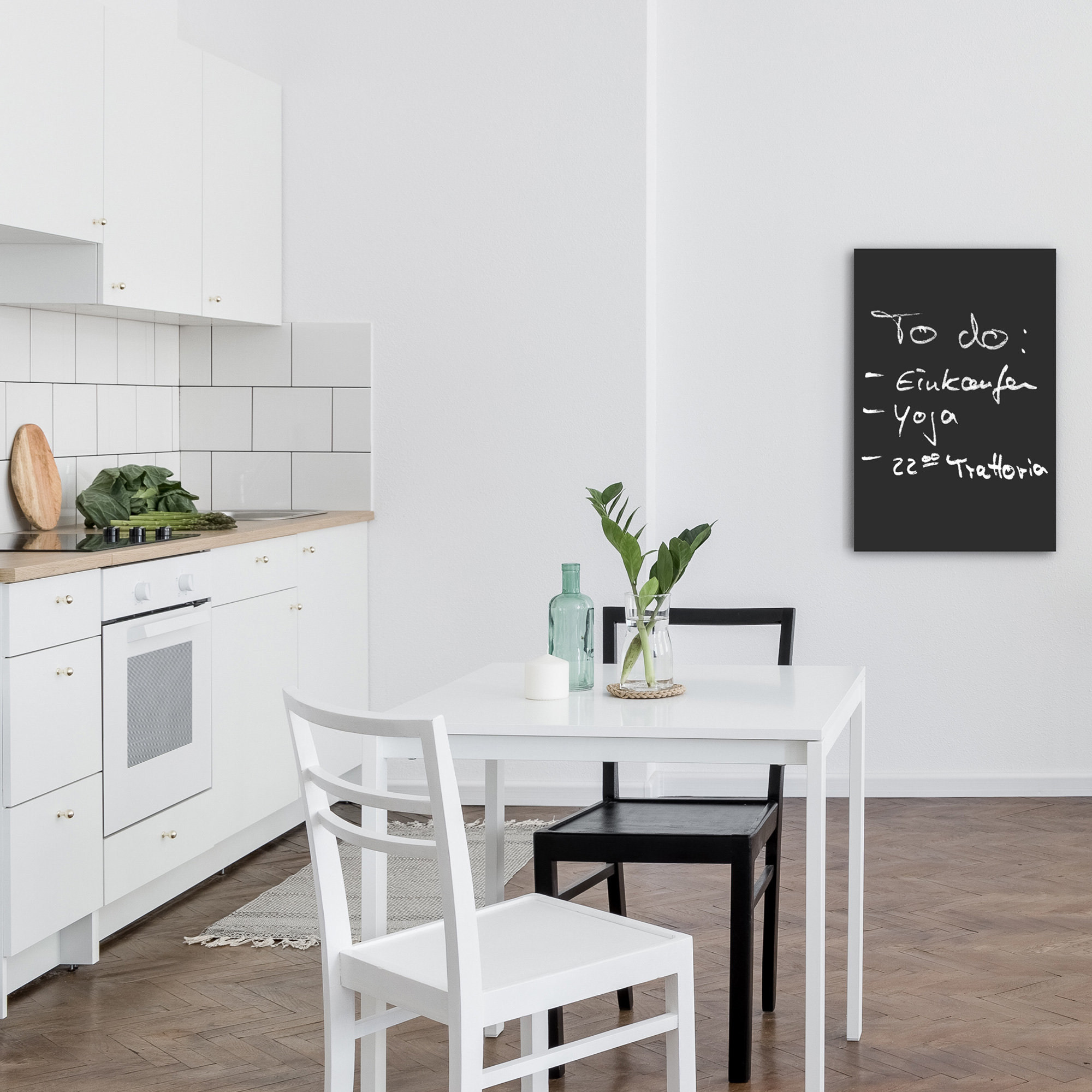 Weiße Küche mit Tisch, zwei Stühlen und einer Tafel mit einer To-Do-Liste. Eine Vase steht auf dem Tisch.