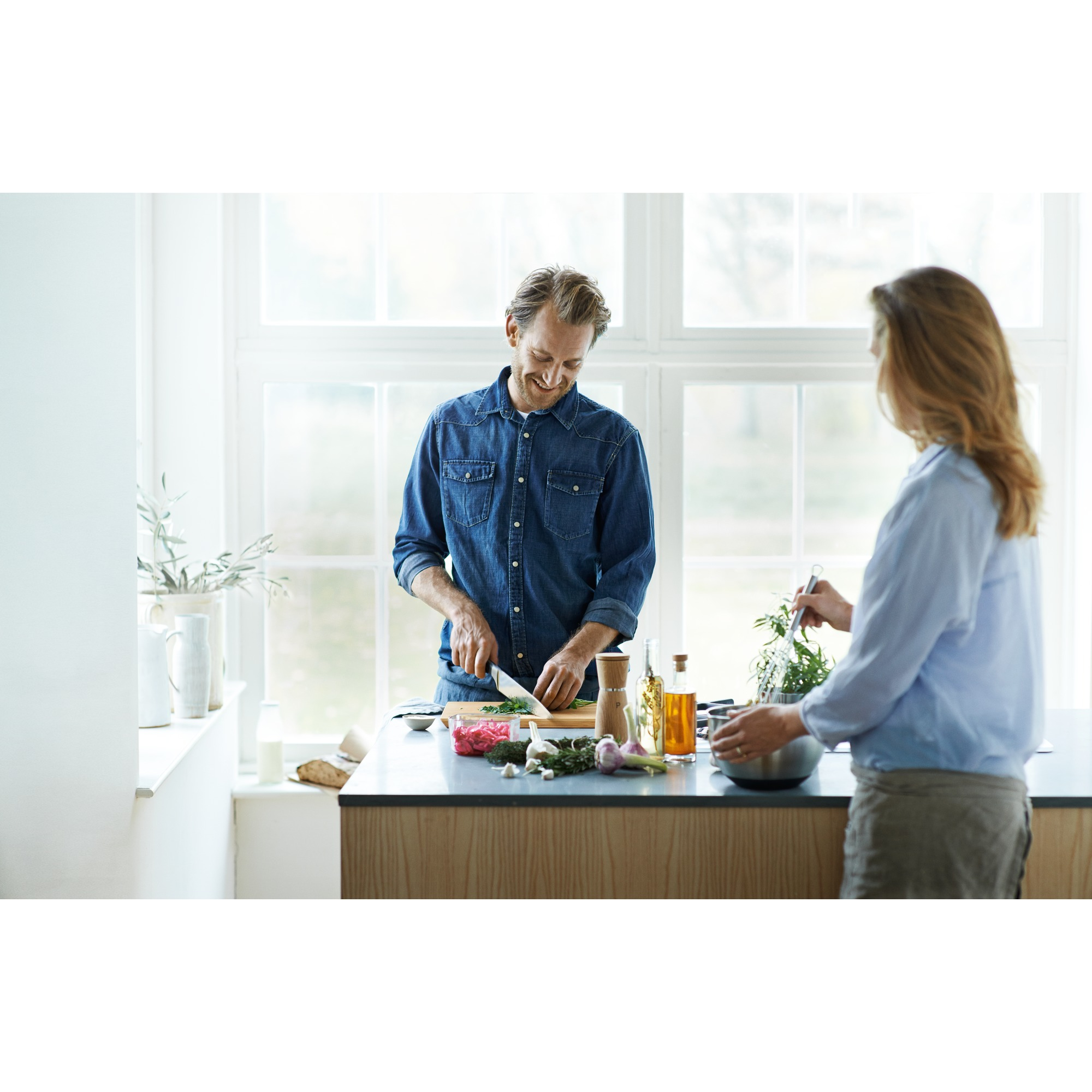 Dwie osoby gotują w jasnej kuchni. Mężczyzna kroi warzywa, kobieta miesza w misce. Białe okno.