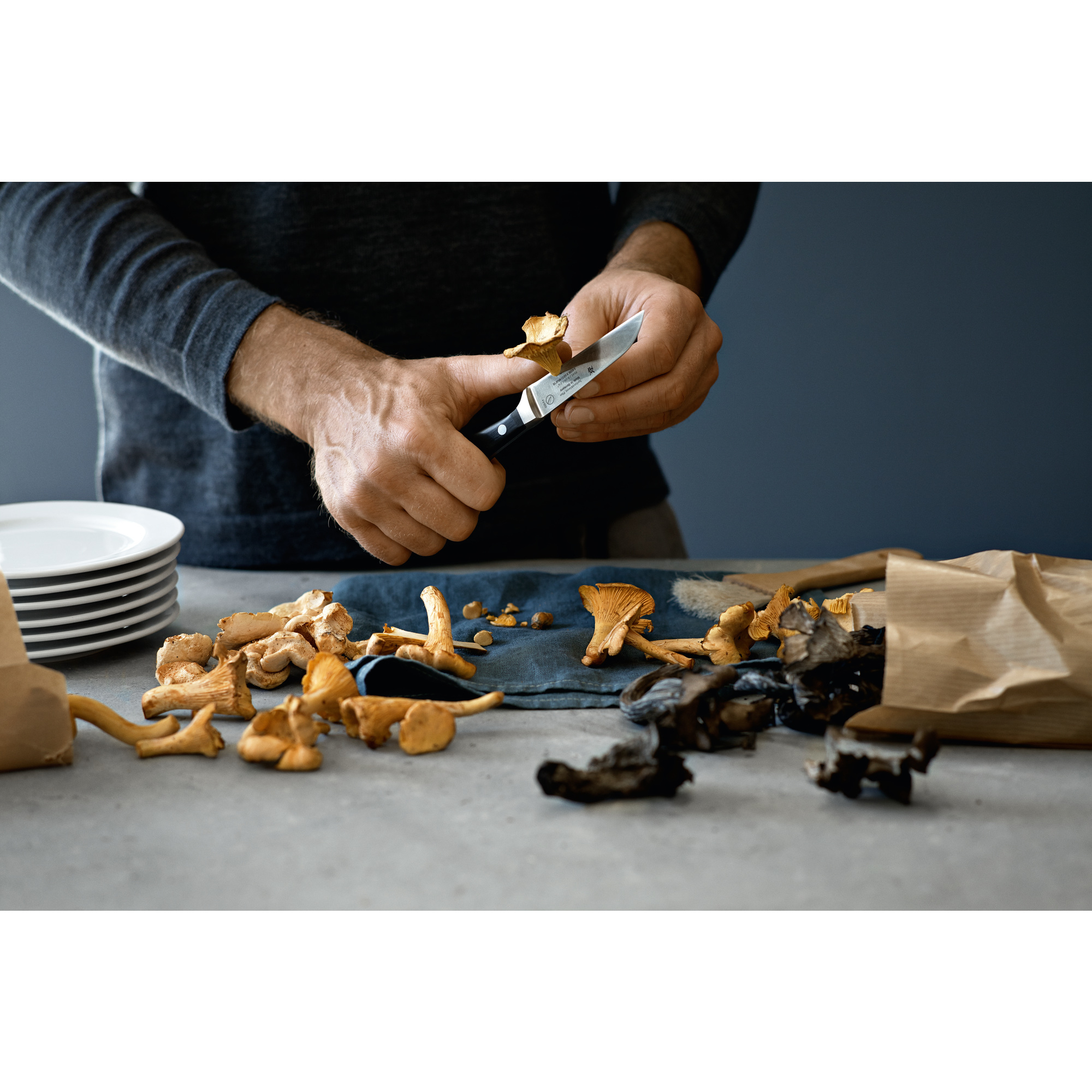 Osoba kroi grzyby nożem na stole, z innymi grzybami i talerzami.