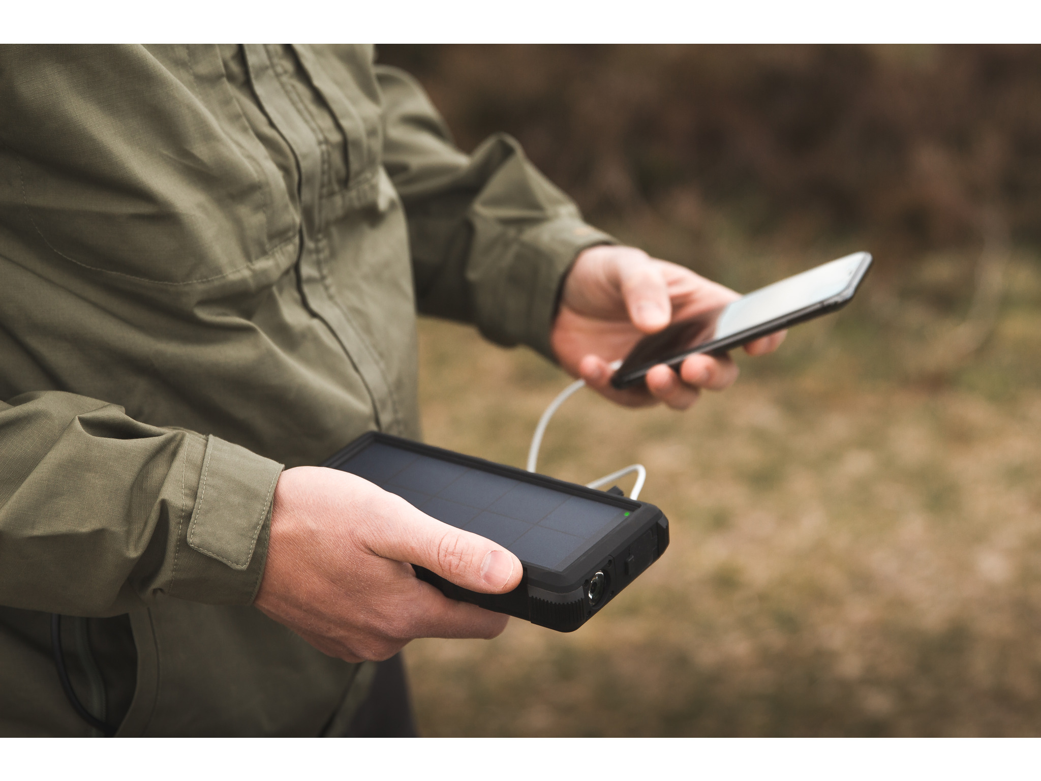 Una persona tiene in mano un telefono e un power bank solare nero, con uno sfondo sfocato all'aperto.