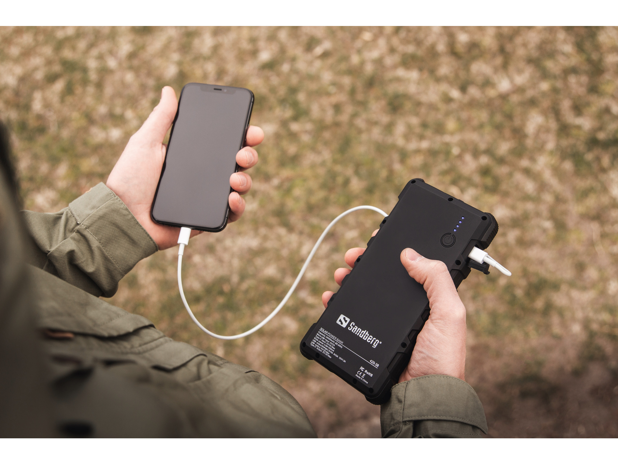 Mani che tengono un telefono e un power bank nero con cavo bianco. Sfondo: erba verde sfocata.
