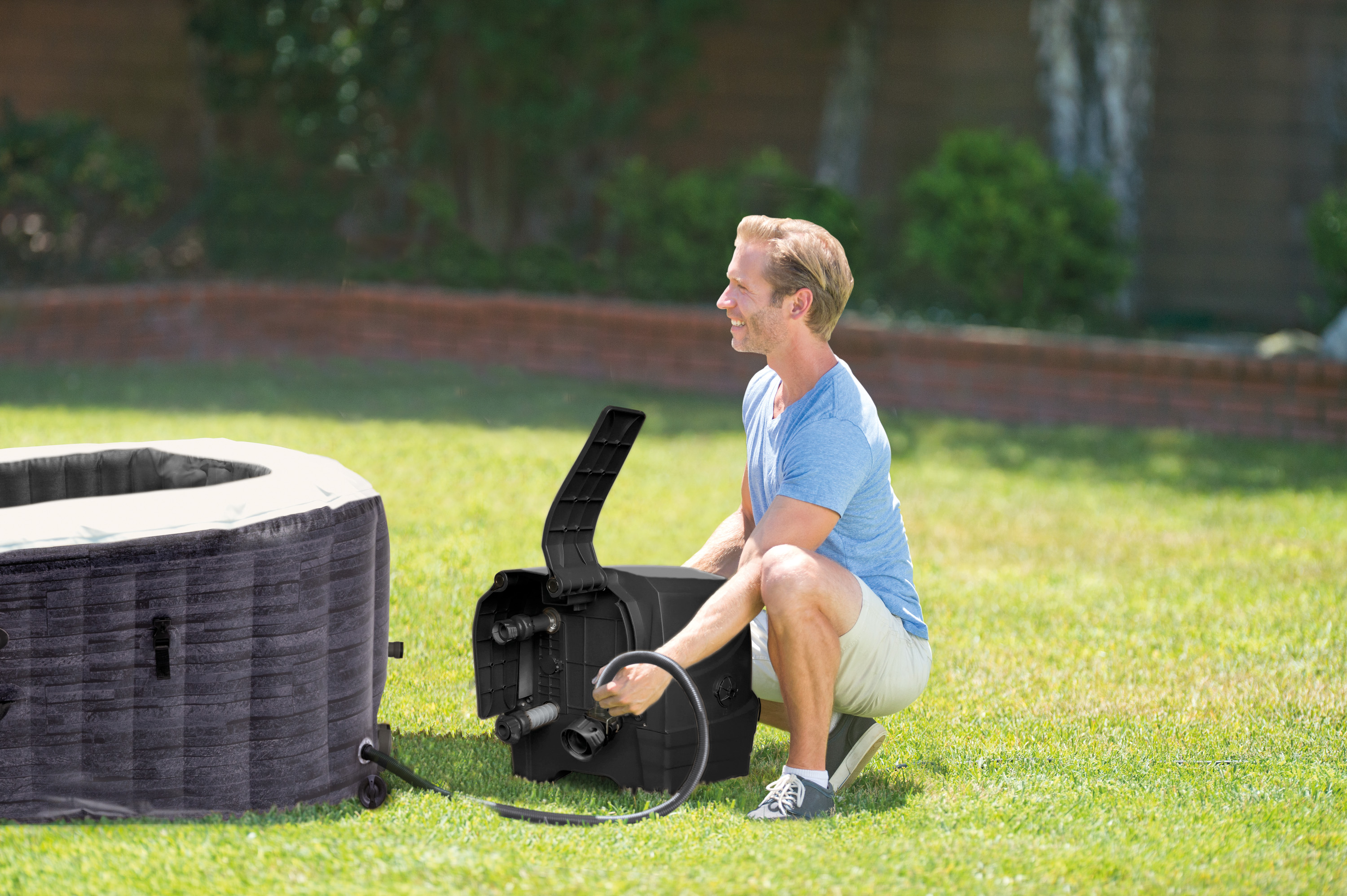 Mężczyzna w niebieskiej koszuli obok czarnej nadmuchiwanej wanny z hydromasażem na trawie. Słoneczny dzień.