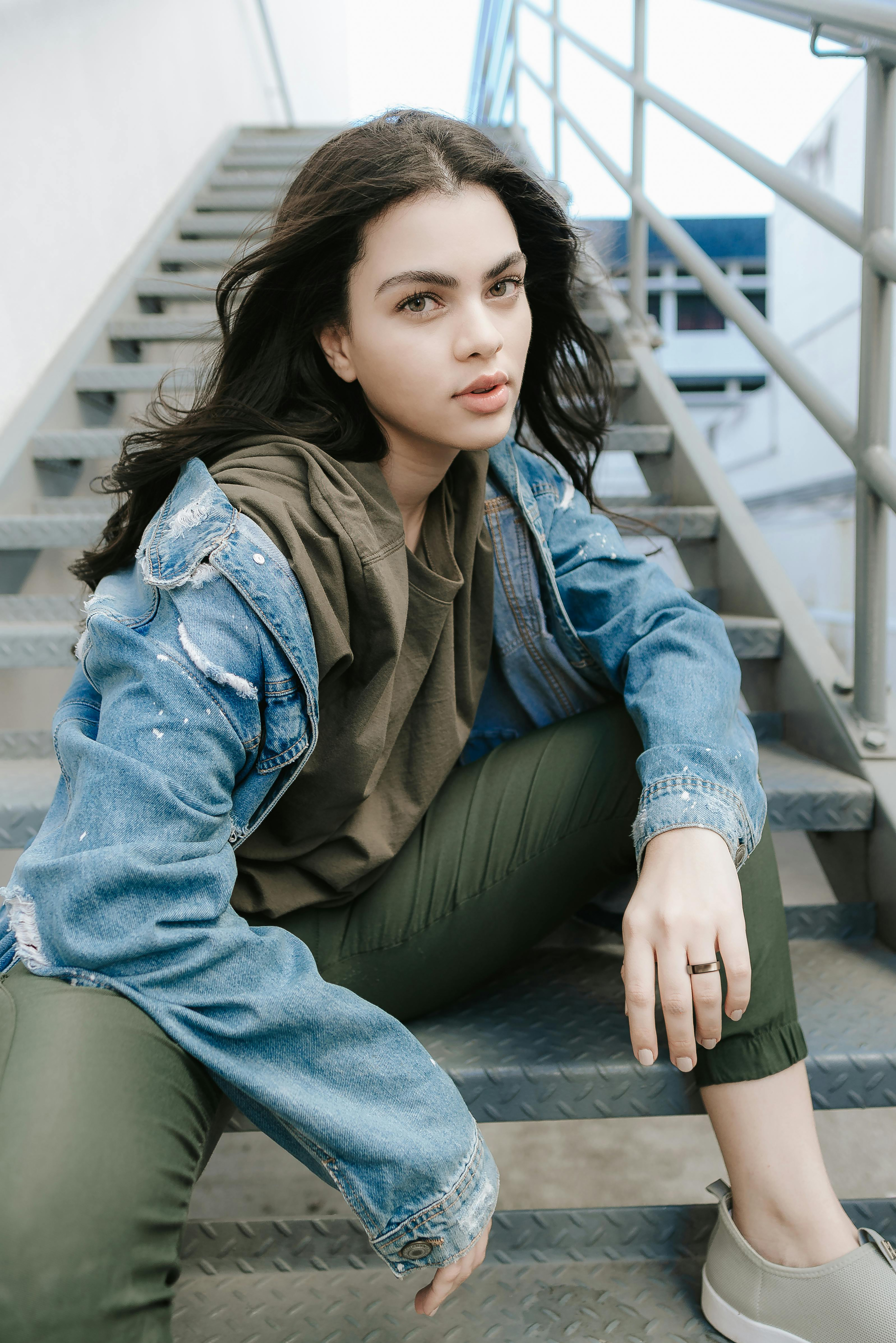 Junge Frau sitzt auf einer Metalltreppe und trägt eine Jeansjacke und olivgrüne Kleidung. Der Hintergrund ist ein Gebäude.