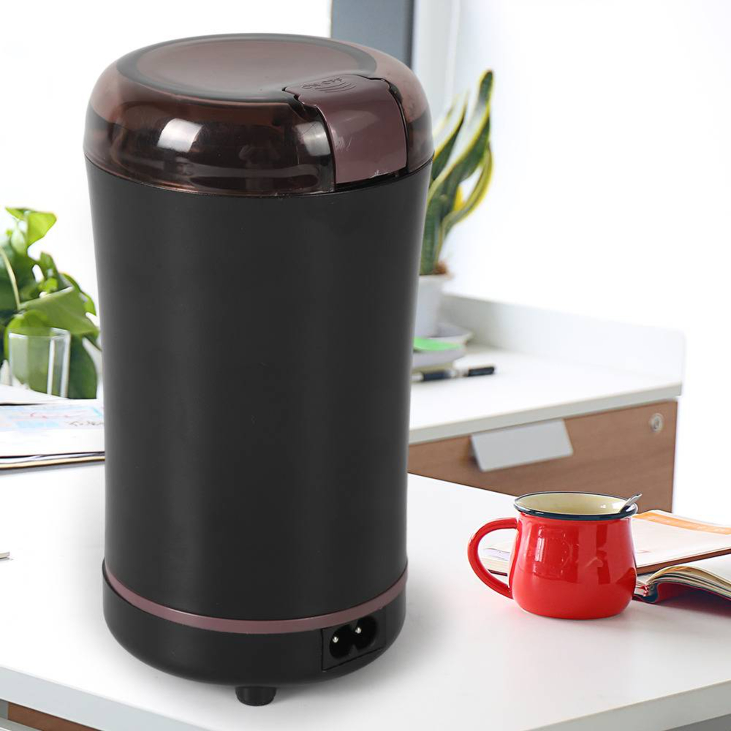 Schwarze Kaffeemühle mit braunem Deckel, roter Becher und offenes Buch auf weißer Oberfläche.