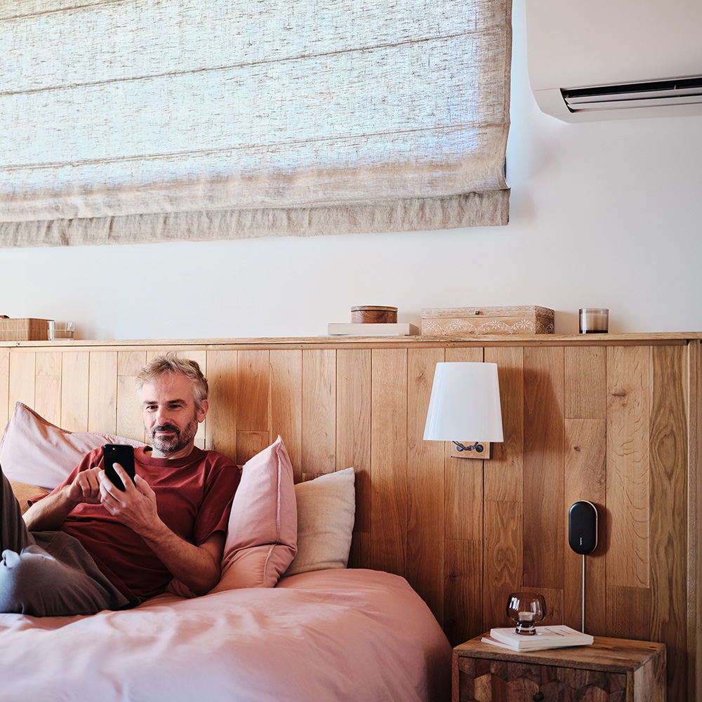 Man in bed with a phone, interior with wooden headboard and lamp.