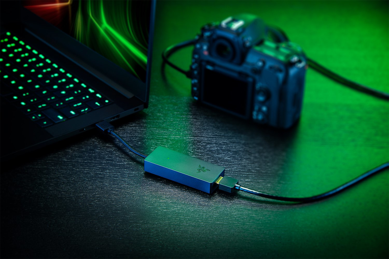 Laptop, fotocamera e un dispositivo Razer blu collegati con cavi, su una superficie scura.