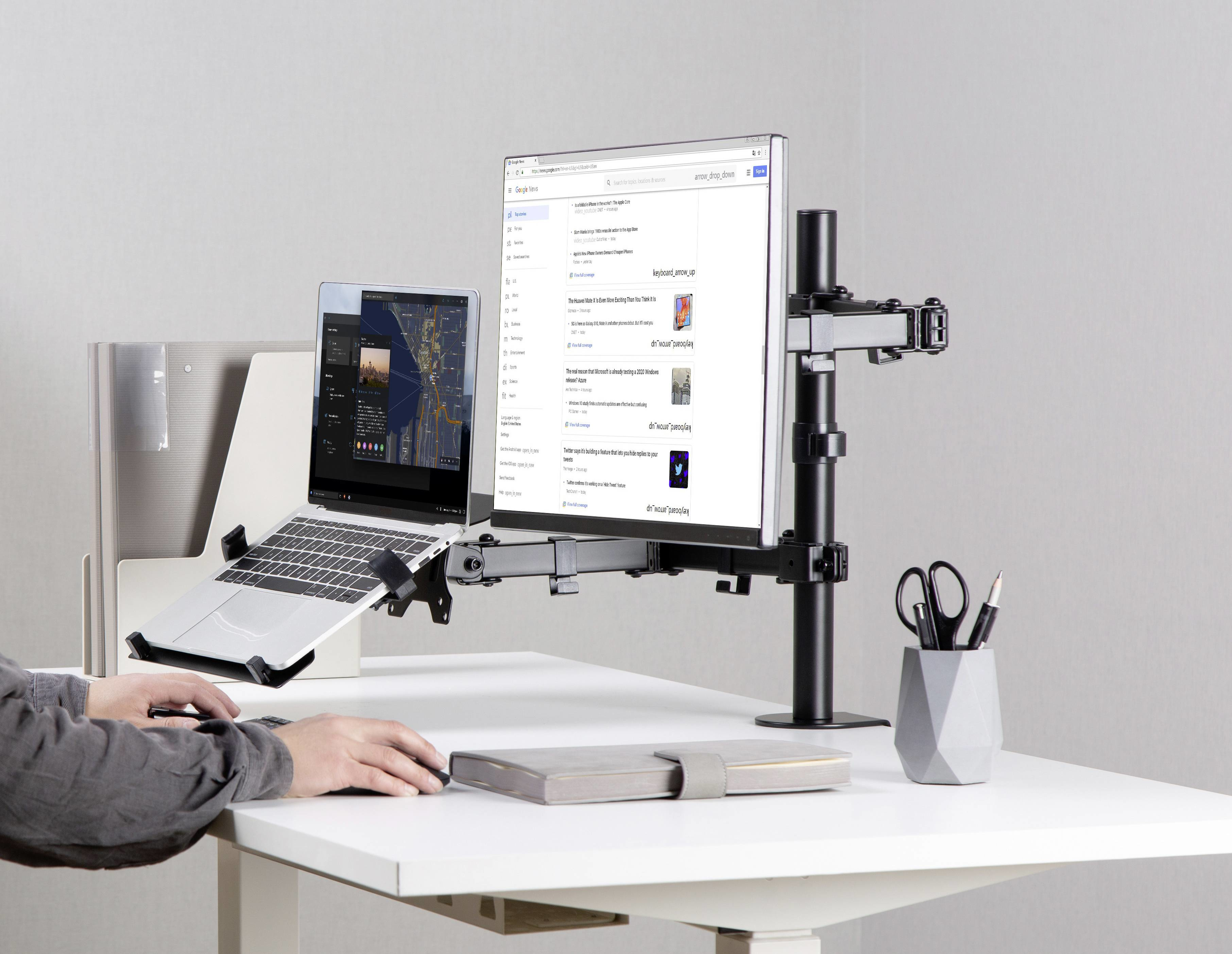 Configurazione scrivania con laptop, monitor e braccio monitor. Mani sulla tastiera, oggetti sulla scrivania in vista.