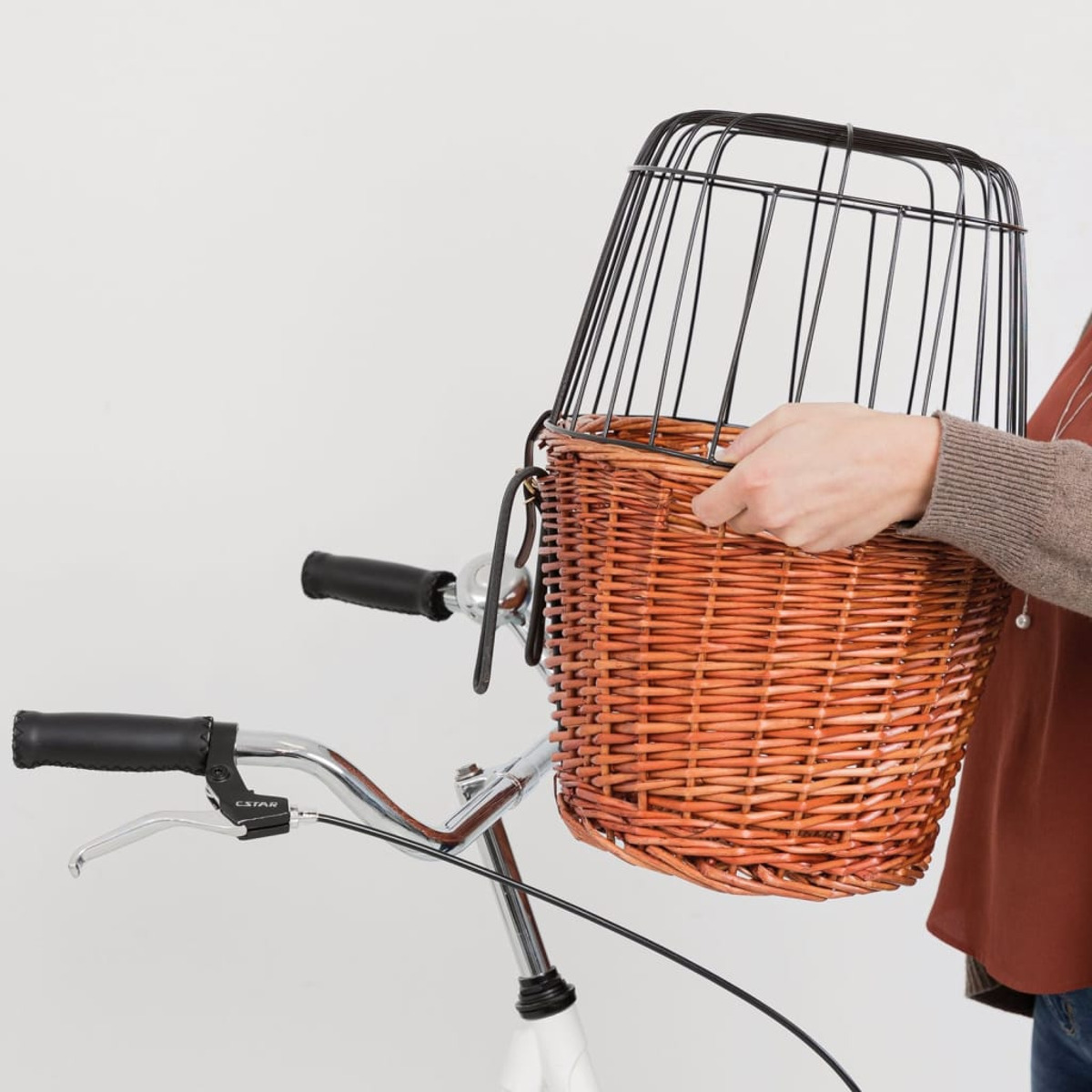Koszyk rowerowy trzymany jest w pobliżu kierownicy białego roweru. Koszyk jest z brązowej wikliny.