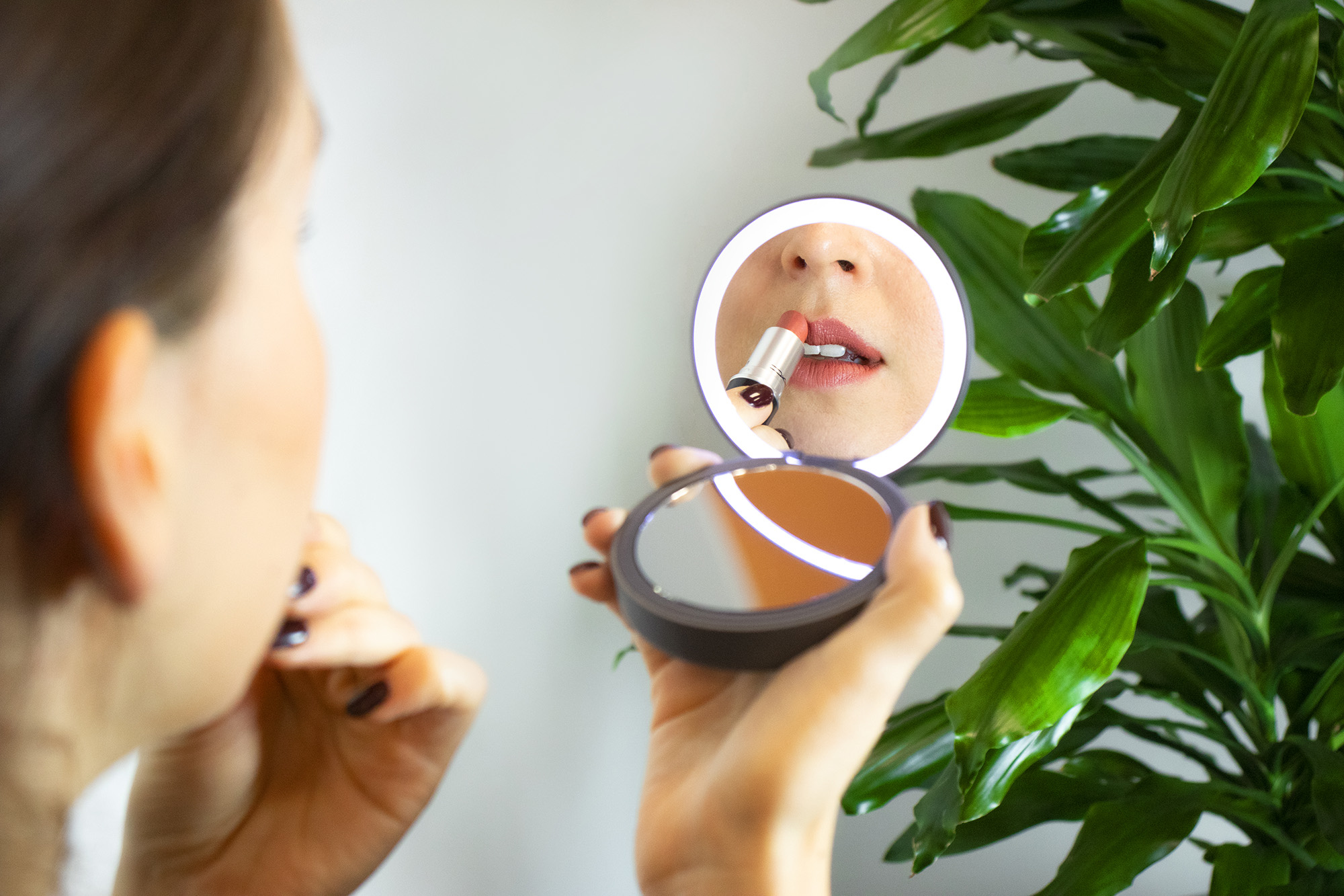 Frau trägt mit einem kleinen Spiegel Lippenstift auf. Grüne Pflanze im Hintergrund. Spiegel reflektiert Gesicht und Lippenstift.
