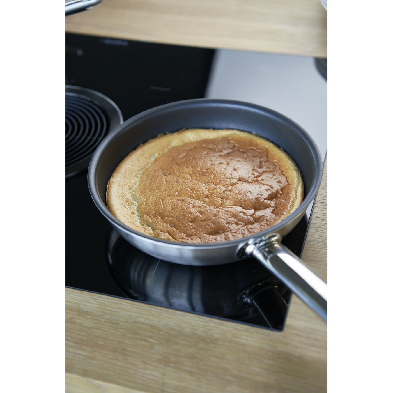 Ein runder Kuchen in einer Metallpfanne auf einem schwarzen Herd, mit Griff rechts.