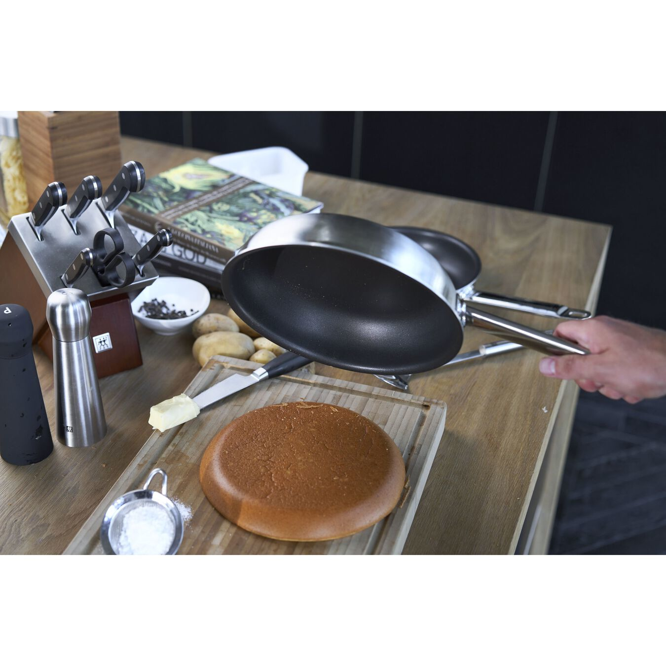 Person hält eine Pfanne über einer Holzoberfläche mit Essen, Messern und Zutaten.