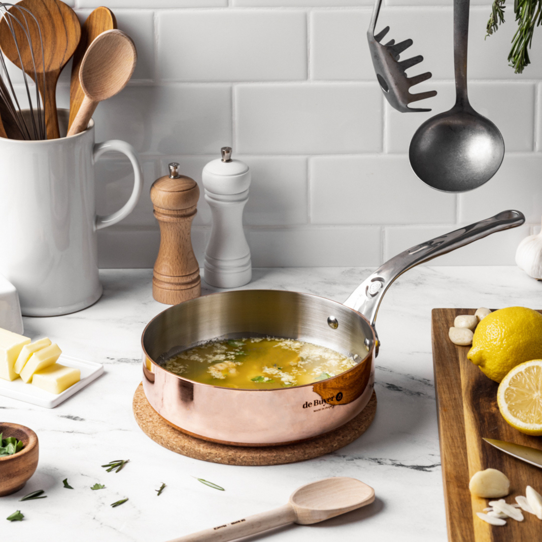 Miedziany rondel na podkładce korkowej, otoczony składnikami na marmurowej powierzchni.