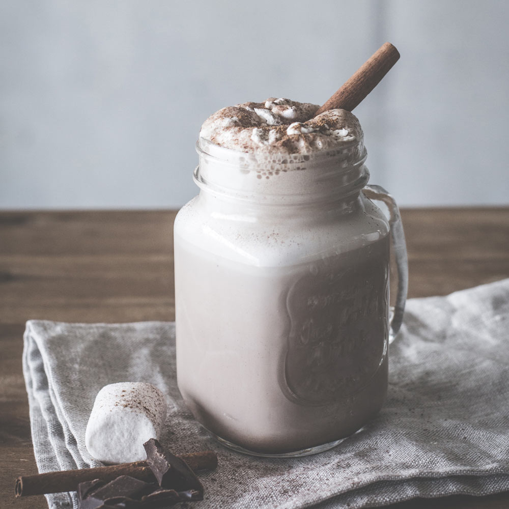 Ein Einmachglas gefüllt mit heißer Schokolade, bedeckt mit Sahne und einem Zimtstange. Ein Marshmallow daneben.