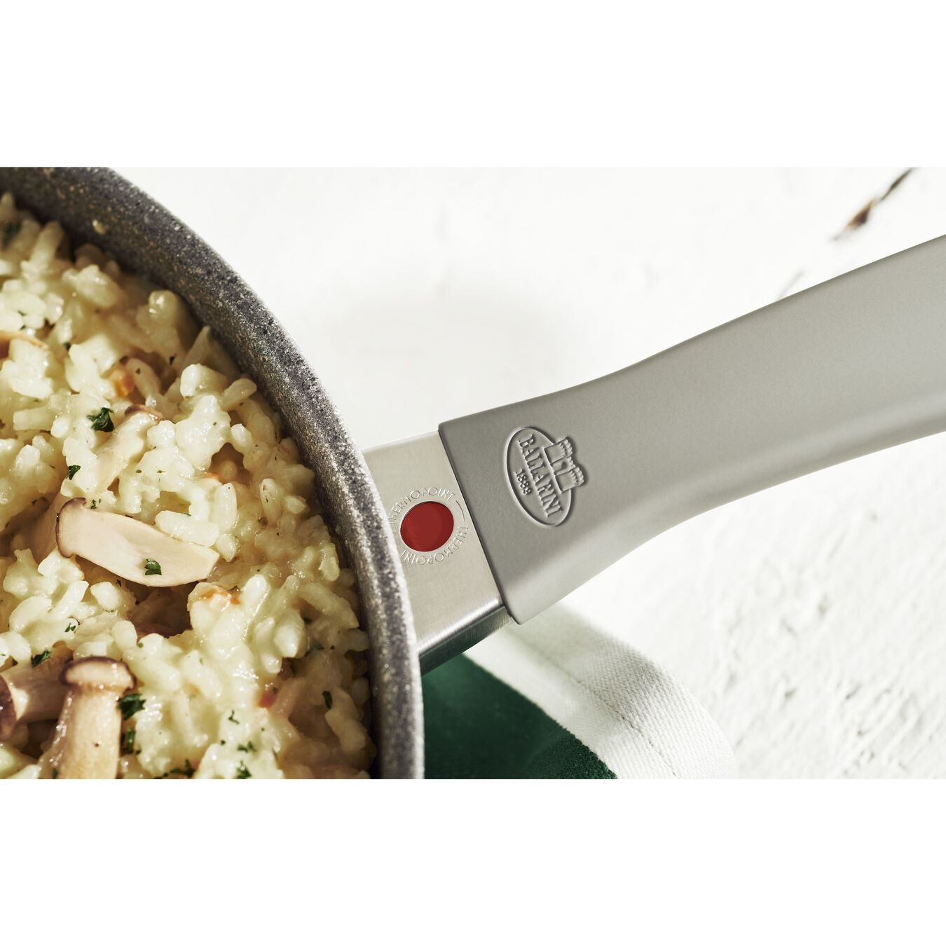 Zbliżenie uchwytu patelni z logo 'Ballarini', z risotto w patelni.
