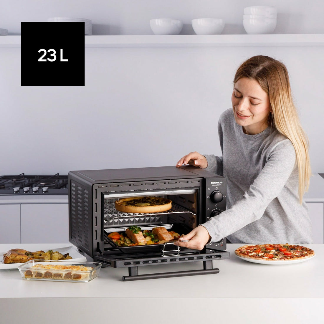 Una mujer usa un horno de 23L para cocinar alimentos. Hay platos cocinados y una pizza en la mesa.