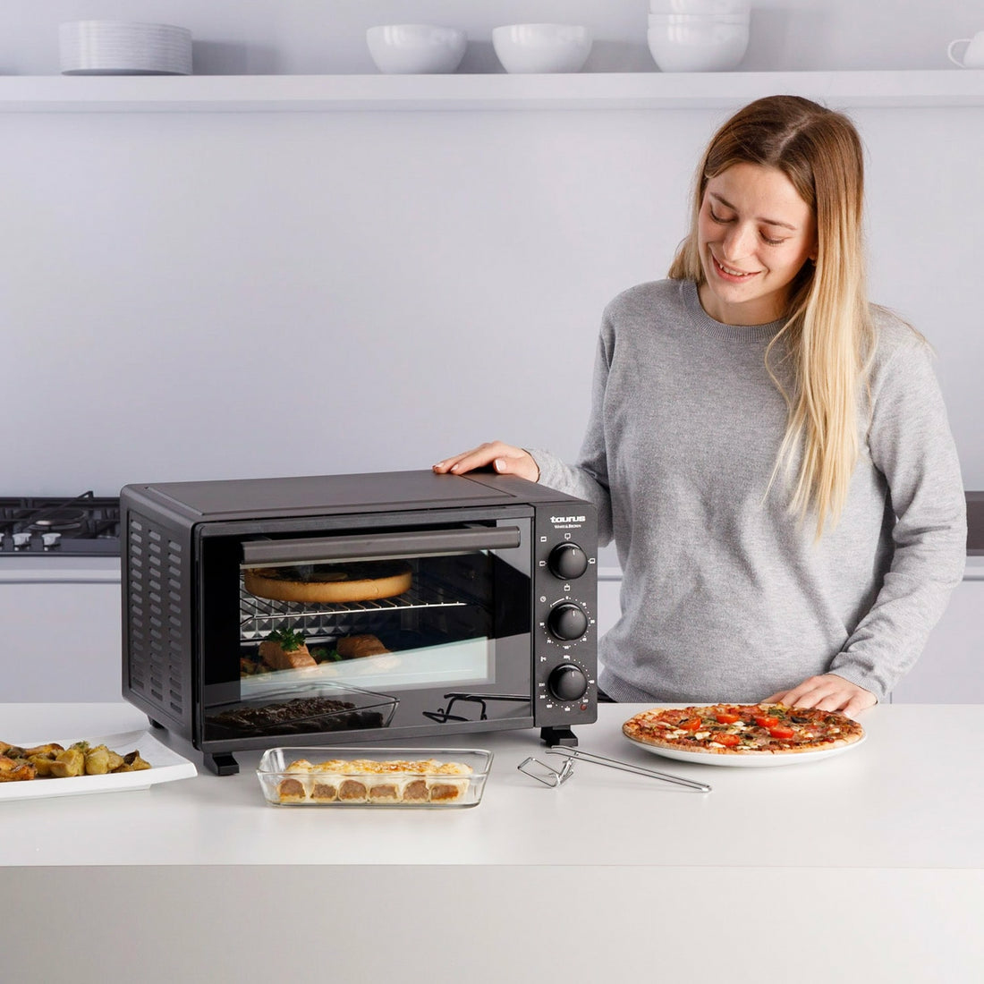 Una mujer sonríe junto a un horno negro con pizza y otros alimentos en la encimera. Fondo de cocina.