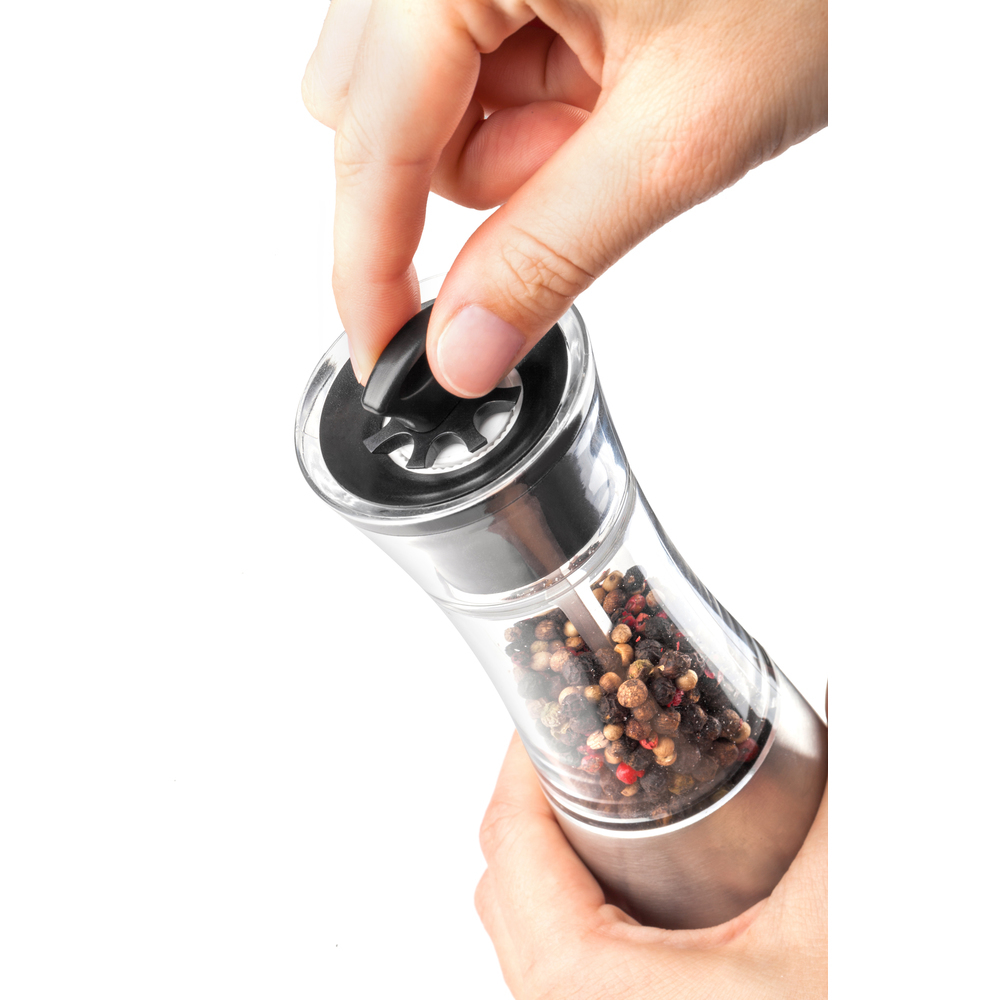 Zbliżenie dłoni uzupełniającej młynek do pieprzu ziarnami pieprzu. Przezroczysty pojemnik, podstawa ze stali nierdzewnej. Białe tło.