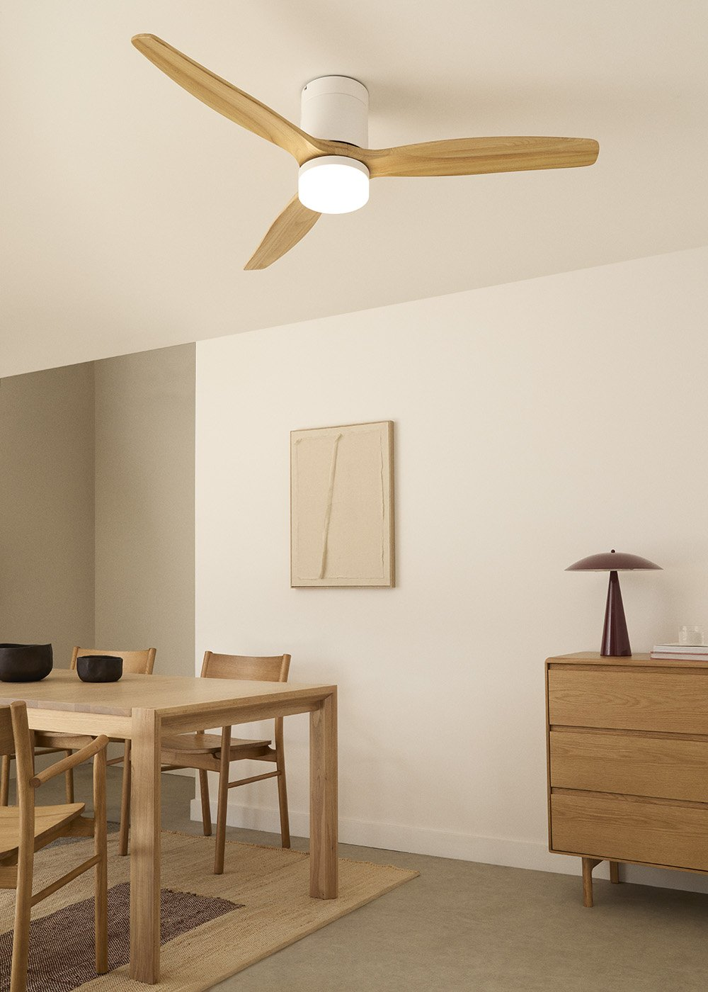 Una habitación con ventilador de techo y mesa con sillas. Una pintura y una cómoda en la pared. Tonos neutros.